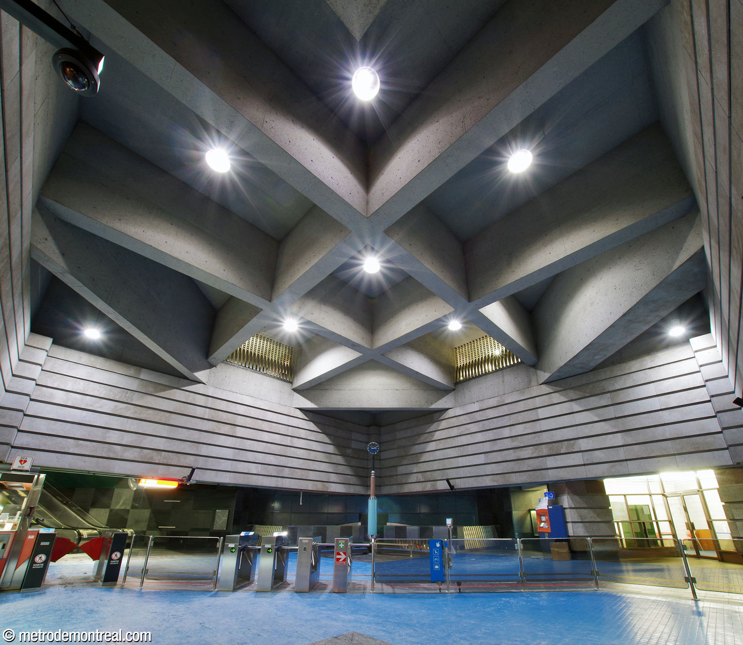 The ticket hall mezzanine, looking southward from the head of the platform stairs toward the gateline. The upper part of the volume is clad in pale grey limestone panels and the ceiling presents a grid of square coffers. At top centre, two openings look down on the ticket hall from the walkway between the headhouses. Below them is Lieu de rencontre, the sculptural tall clock by Météore Design.