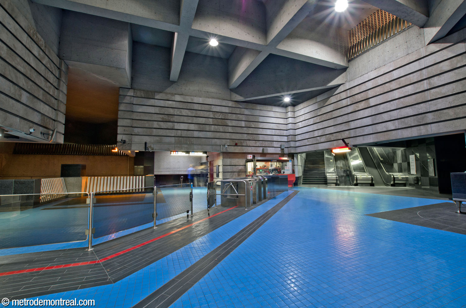 The ticket hall, looking east along the gateline toward the ticket booth. The ticket hall's shape as a square set at a 45-degree angle can be clearly seen, and is emphasized by the bold design of the azure and black floor tiles. At right is the stairwell leading to the exits. At centre-left are the stairs to the Saint-Michel platform, and at left, the overlook onto the platforms.