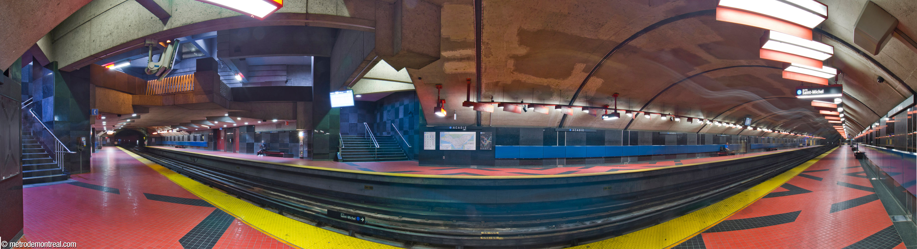 Panorama of the trainroom at Acadie station, with the mezzanine and stairs up to it at left. The platform and stairwell walls are a checkerboard of matte and polished black granite, with a stripe of dark blue panels along the platform. The floors are bright red-pink tiles with bold black diagonal designs. The platform light fixtures are set at an angle and carry red metal shades on one side. The mezzanine projects out with a prow-like point over the tracks.