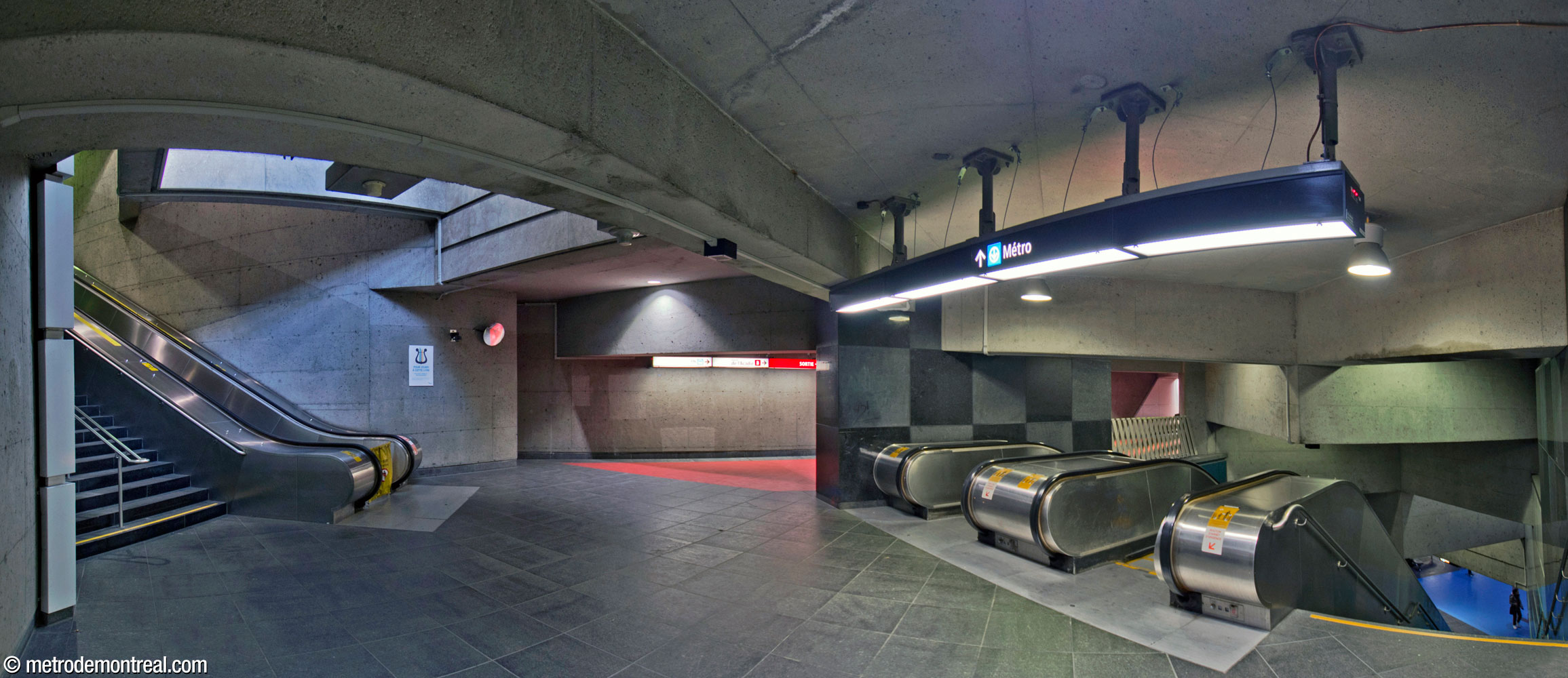 The intersection between the passage between the headhouses (centre) and the stairwell down to the ticket hall (right). At left is the stairwell up to the Beaumont headhouse; a light shaft is located above the foot of the escalator. The passage leads to the De l'Acadie headhouse.