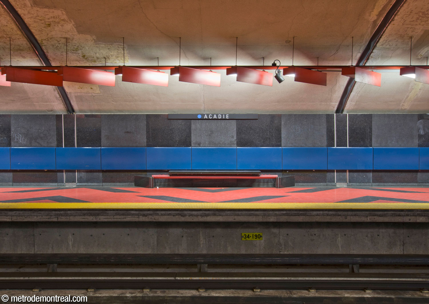 Face-on view of the platform décor. The walls are lined with a checkerboard of polished and matte black granite with a stripe of dark blue enamelled steel panels; the floor is tiled with raspberry red and black tiles. We also see the distinctive black granite and red steel benches and the unique effect produced by the diagonally set light fixtures above.