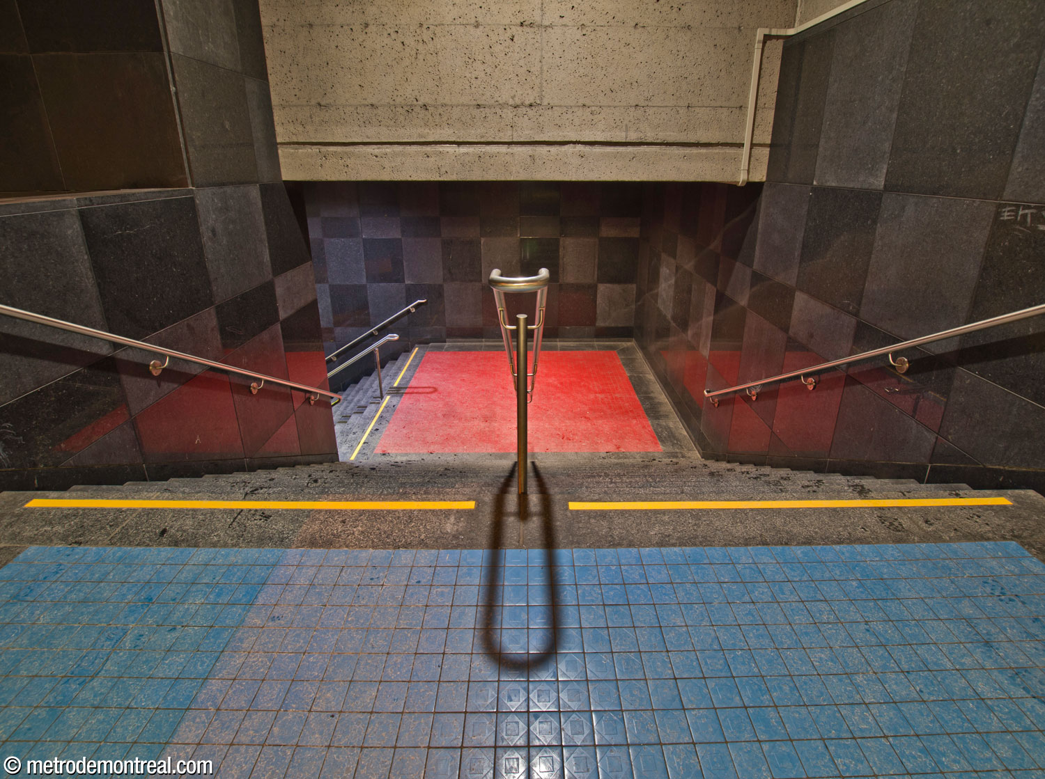 Looking down the stairs toward the Saint-Michel platform. At the head of the stairs, we see the azure floor tiles of the mezzanine, and on the landing, the red tiles of the platforms. The walls of the stairwell are clad in the checkerboard of matte and polished black granite used on the platform walls.
