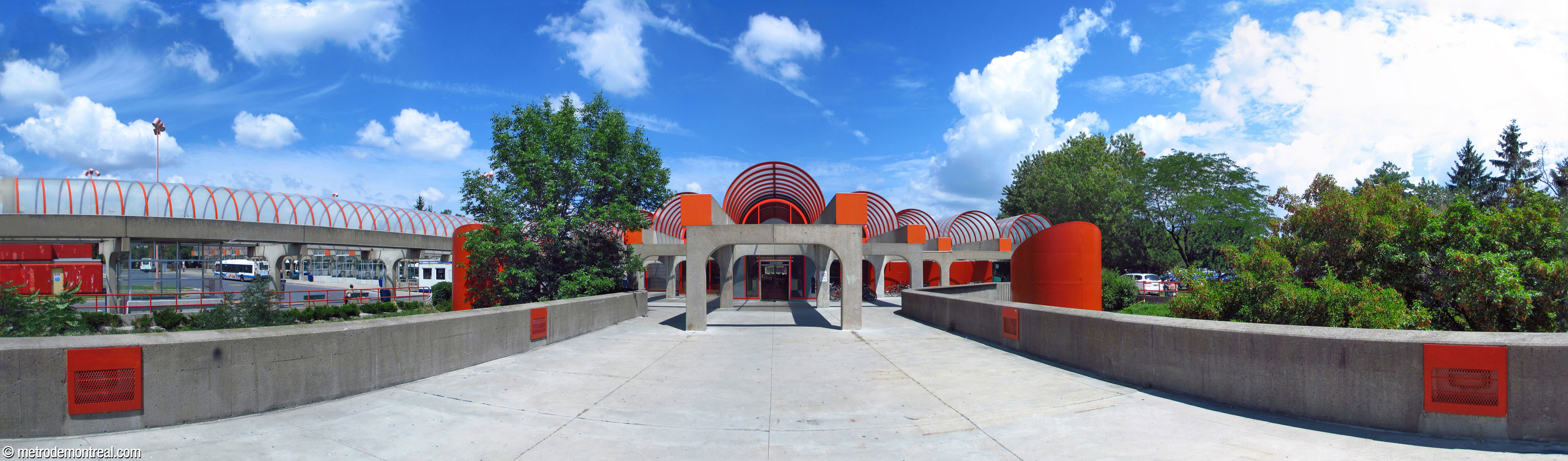 Exterior of the headhouse, seen from the southwest side and the walkway leading from Parc Angrignon. The building's scaffolding-like framework extends outward from its walls, bearing its semi-cylindrical roof vaults. The orange funnels on either side are ventilation shafts. The bus terminus is located at left.