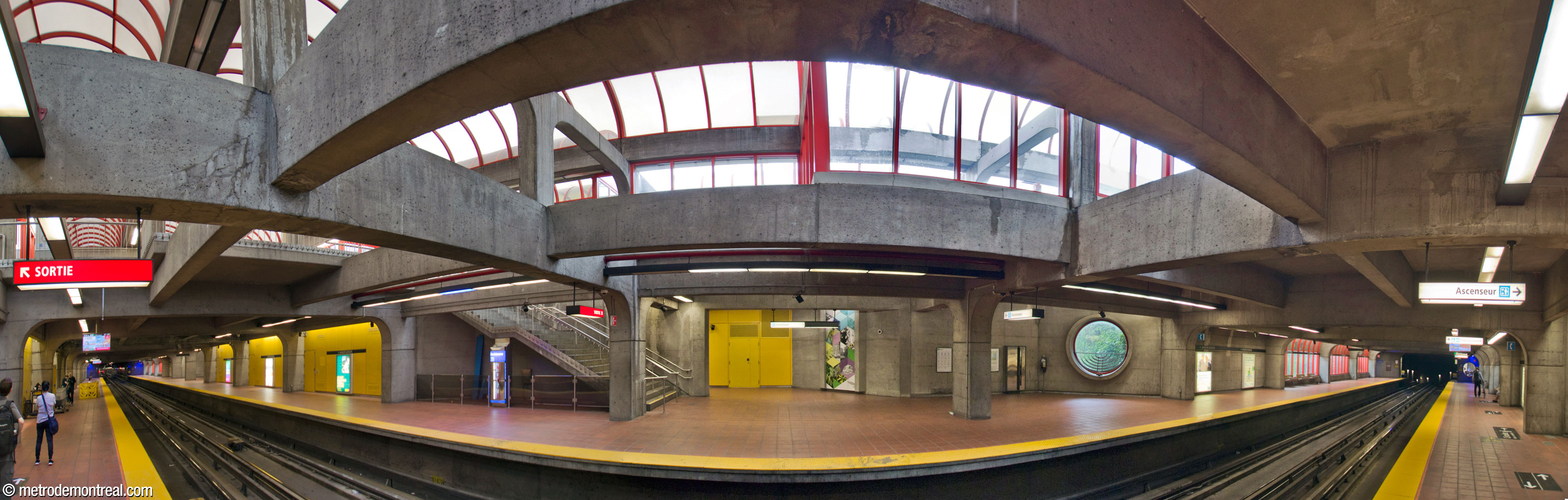 Panorama of the station platforms, seen from the foot of the stairs to the departure platform. The ends of the platforms receive sunlight from platformside windows, while the central area is lit by the transparent domes overhead. The brightness is enhanced by bright yellow panels on the walls. One section of Shelley Miller's artwork Les boîtes vivantes is seen on the opposite platform.
