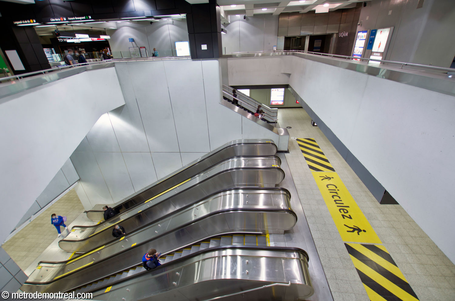 Un large volume au-dessus des escaliers entre le niveau de la billetterie et le quai Côte-Vertu, avec les escaliers mécaniques qui continuent jusqu'au quai Honoré-Beaugrand. En haut au fond, le couloir vers la sortie Sainte-Catherine (gauche) et l'accès au pavillon Judith-Jasmin (droite).