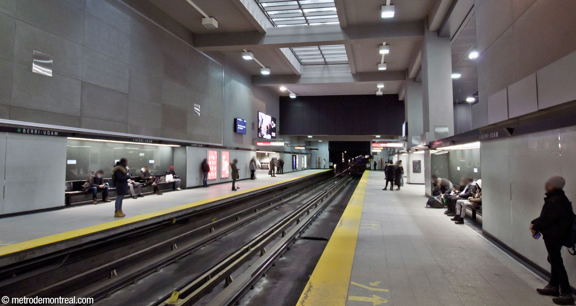 Autre vue de la partie ouest des quais de la ligne verte, montrant les finis neutres utilisés à travers la station : pastilles de pâte de verre blanche en bas, béton lisse en haut, avec des panneaux de verre gris et des accents noirs.