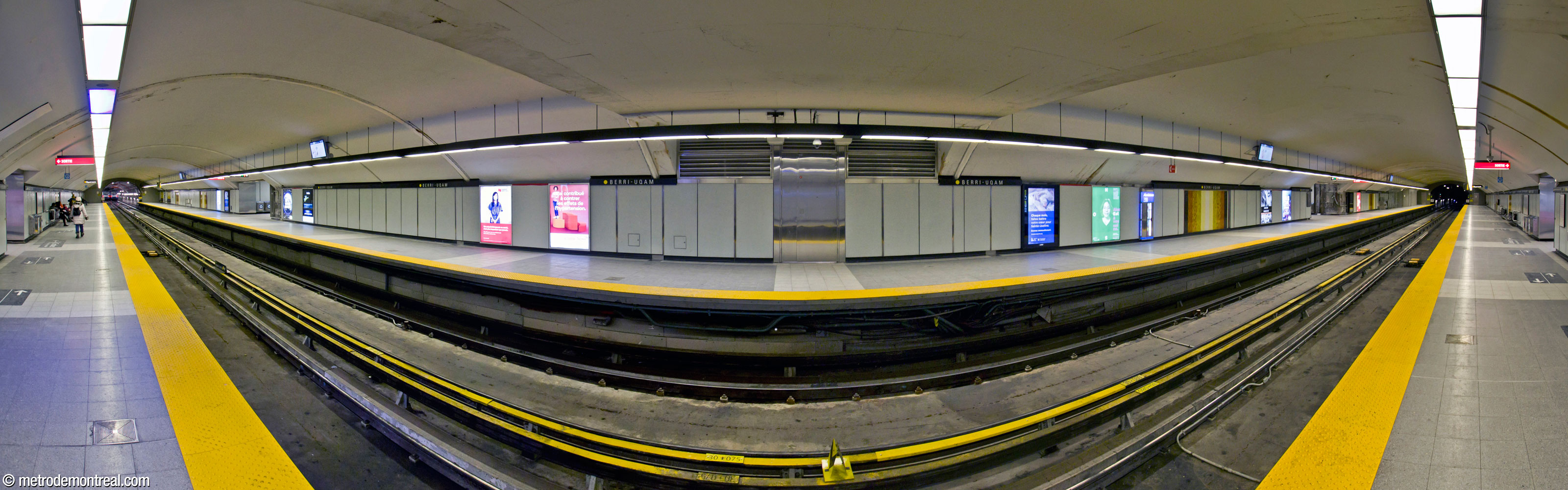 Panorama des quais de la ligne jaune, vus du quai de départ. Le plafond voûté démontre que ce volume a été creusé en tunnel dans le roc, à différence des autres volumes de la station. La section plate du plafond au-dessus du photographe correspond à la passerelle au-dessus de la nef. Des murales carrelées de Claude Vermette égayent les murs.