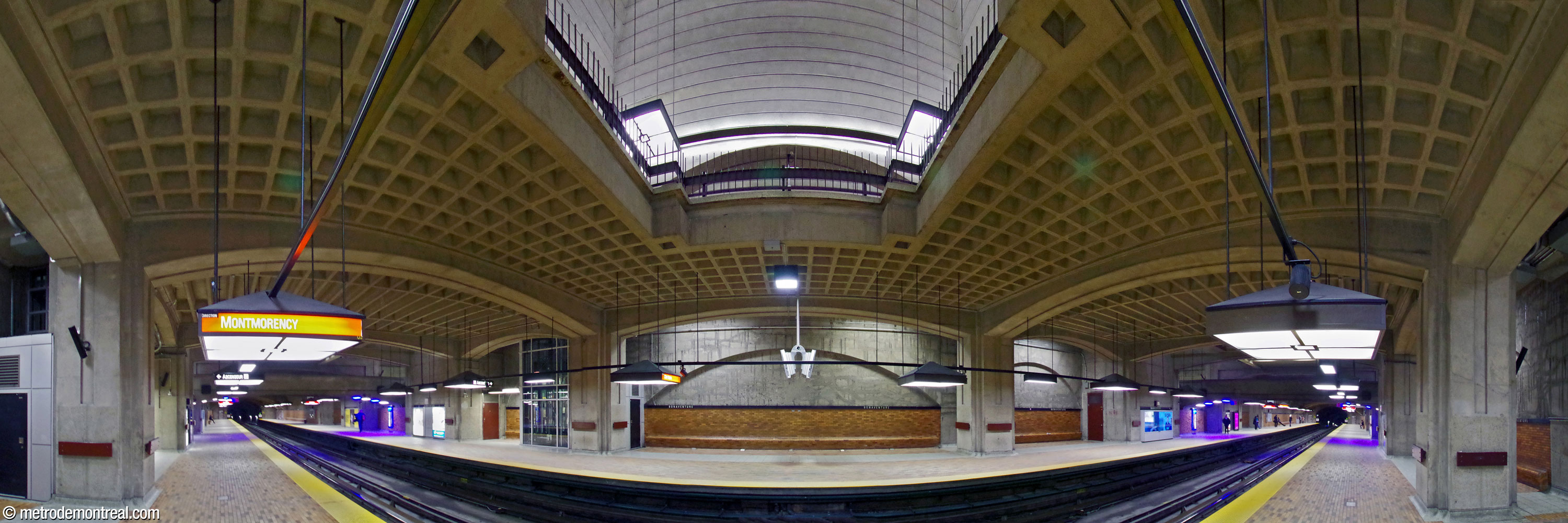Panorama des quais de la station, vus depuis le centre du quai Montmorency. D'imposantes piles soutiennent les voûtes de la station ainsi que la mezzanine qui, elle, forme un plafond à caissons pour les quais. En haut, une percée visuelle à travers la mezzanine offre une vue en contre-plongée des voûtes au niveau supérieur.