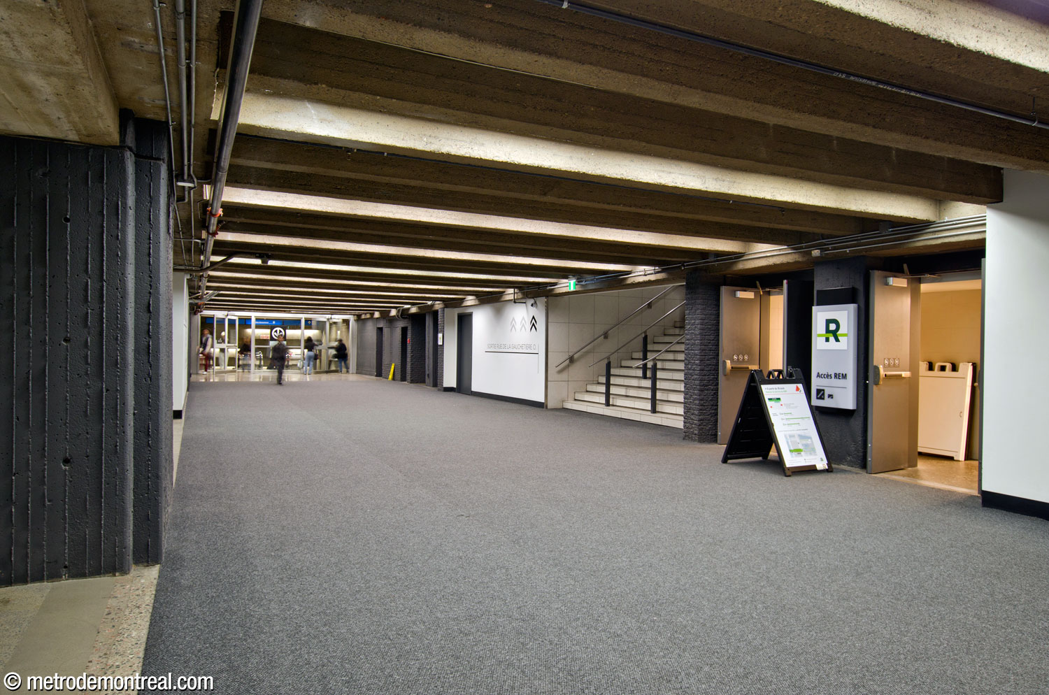 L’escalier à partir du long corridor débouche sur le fond de cette section du sous-sol de la Place Bonaventure. Au moment de l’inauguration du REM, le lien existant de la ville souterraine entre le métro et la gare Centrale s’est avéré plutôt long et tortueux pour supporter les affluences attirées par le nouveau moyen de transport. Or, comme le long corridor débouchait près de l’entrée secondaire de la station du REM (une ancienne sortie à sens unique depuis les quais de la gare Centrale vers la Place Bonaventure), on a pu le recycler pour en faire un lien plus direct vers le REM. Cet espace a été partitionné pour raccorder l’escalier à l’entrée secondaire.