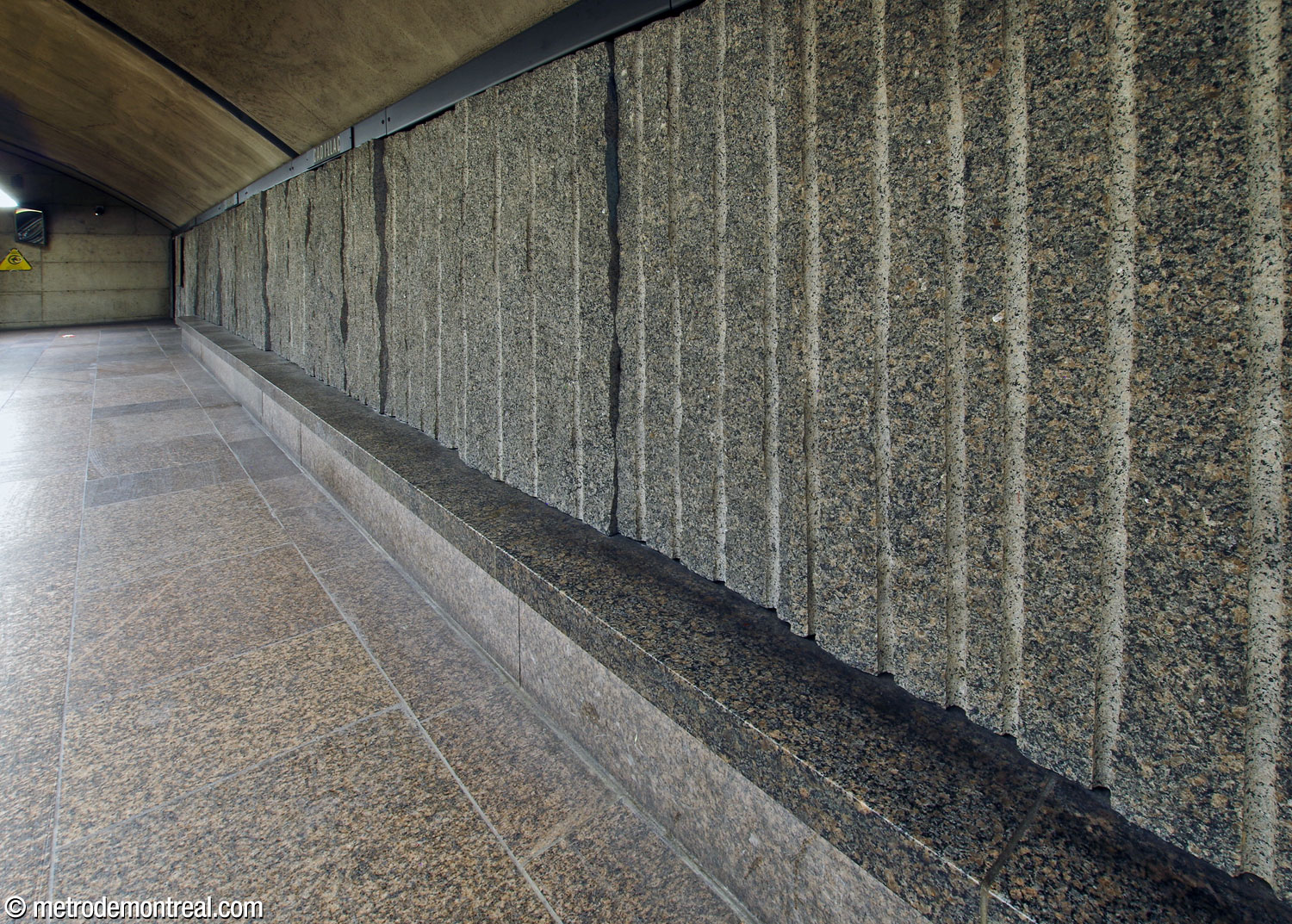 Oblique view of a platform bench, showing the uneven depth of the rough granite wall panels.