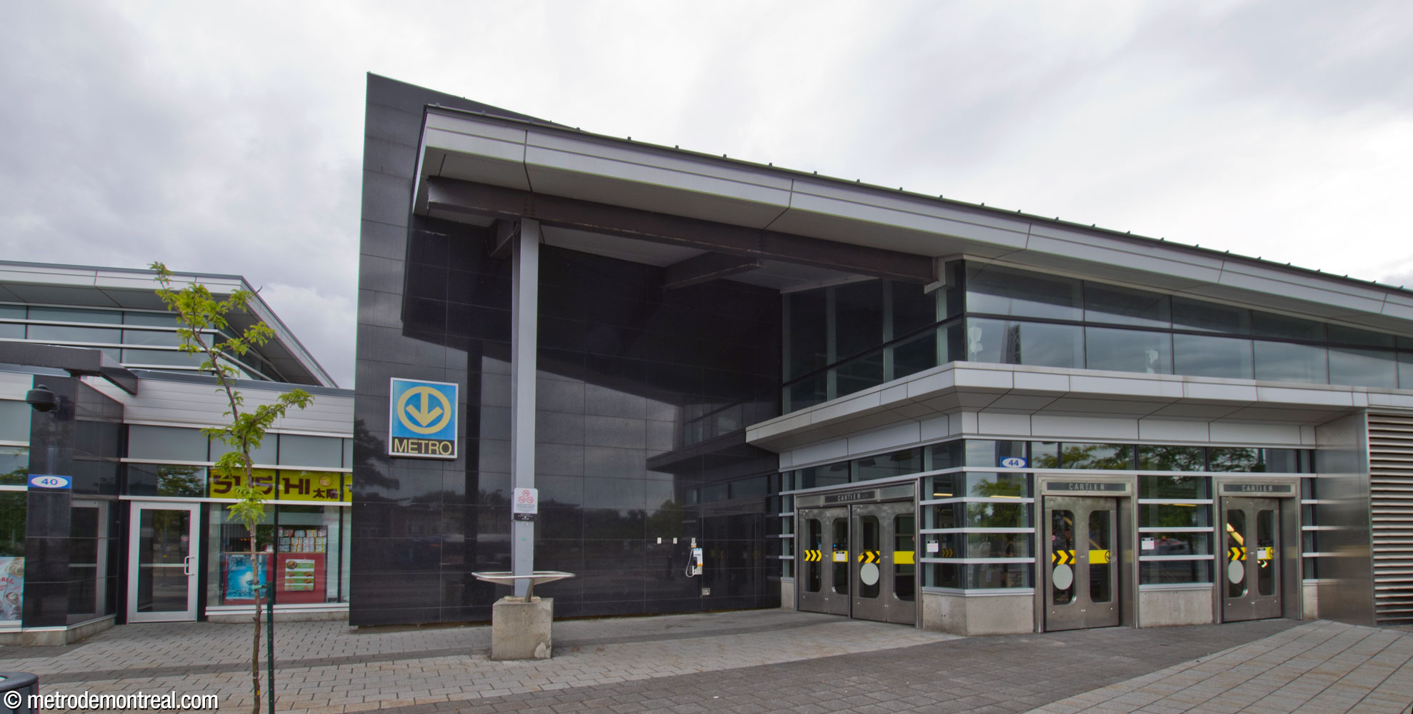 Vue extérieure de l'entrée sud de l'atrium de l'édicule principal. À côté, un plan de granit noir trace une ligne parallèle à celle des voies du métro.