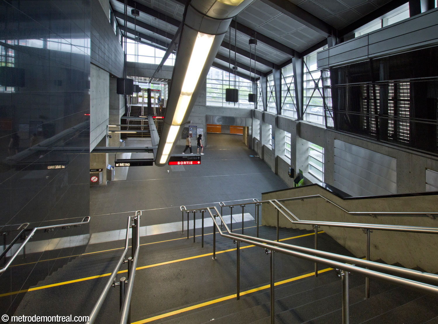 Vue à partir de l'entrée sud de l'édicule principal vers l'atrium surbaissé. À gauche, les escaliers vers la billetterie du métro. Au centre et à droite, des murs de verre font jaillir un éclairage naturel abondant. Au centre droit, l'entrée du tunnel vers l'édicule secondaire.