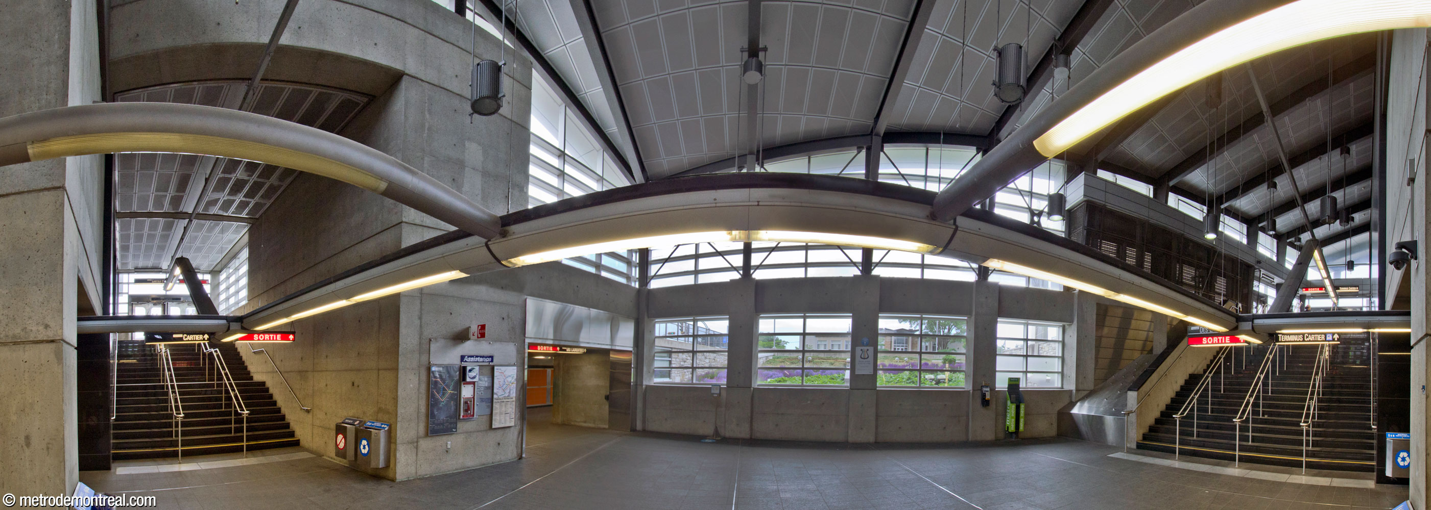 L'atrium de l'édicule principal, vu de la tête des escaliers vers le métro. On voit le jardin surbaissé à l'extérieur à travers le mur vitré qui éclaire la partie centrale. Des escaliers montent vers les sorties nord (gauche) et sud (droite); au centre gauche, un tunnel mène vers l'édicule secondaire.