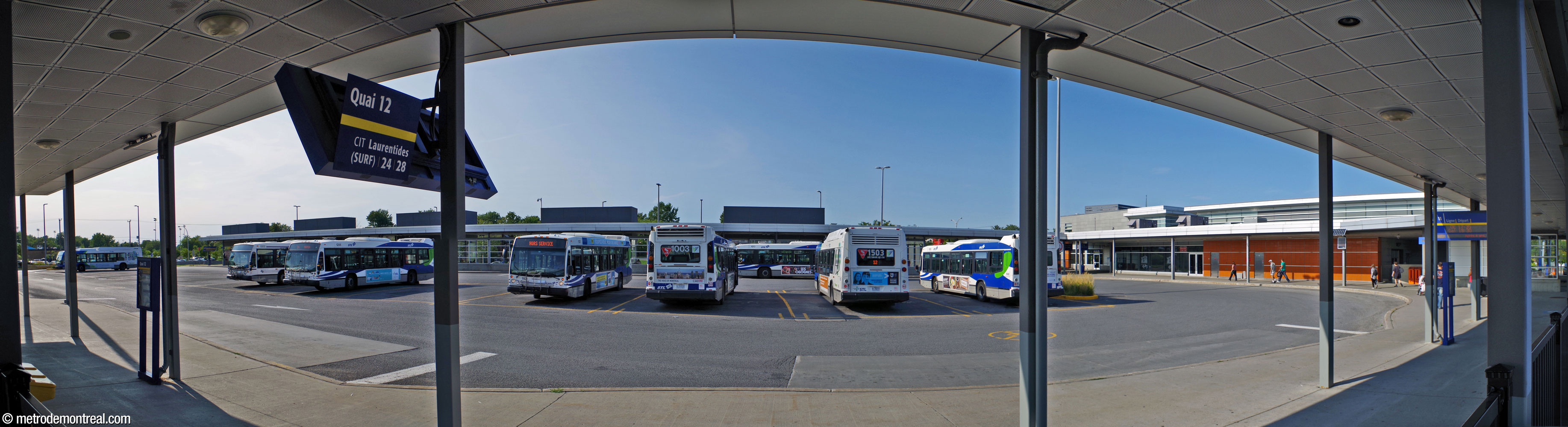 Panorama du terminus d'autobus, vu de sa jetée sud. Les deux jetées se projettent vers l'ouest à partir de l'édicule principal. À droite, des passagers se dirigent vers l'entrée de la partie intérieure du terminus. La terre cuite glacée orange est utilisée plus abondamment sur les murs autour des entrées du terminus d'autobus qu'elle ne l'est sur le devant de l'édicule.