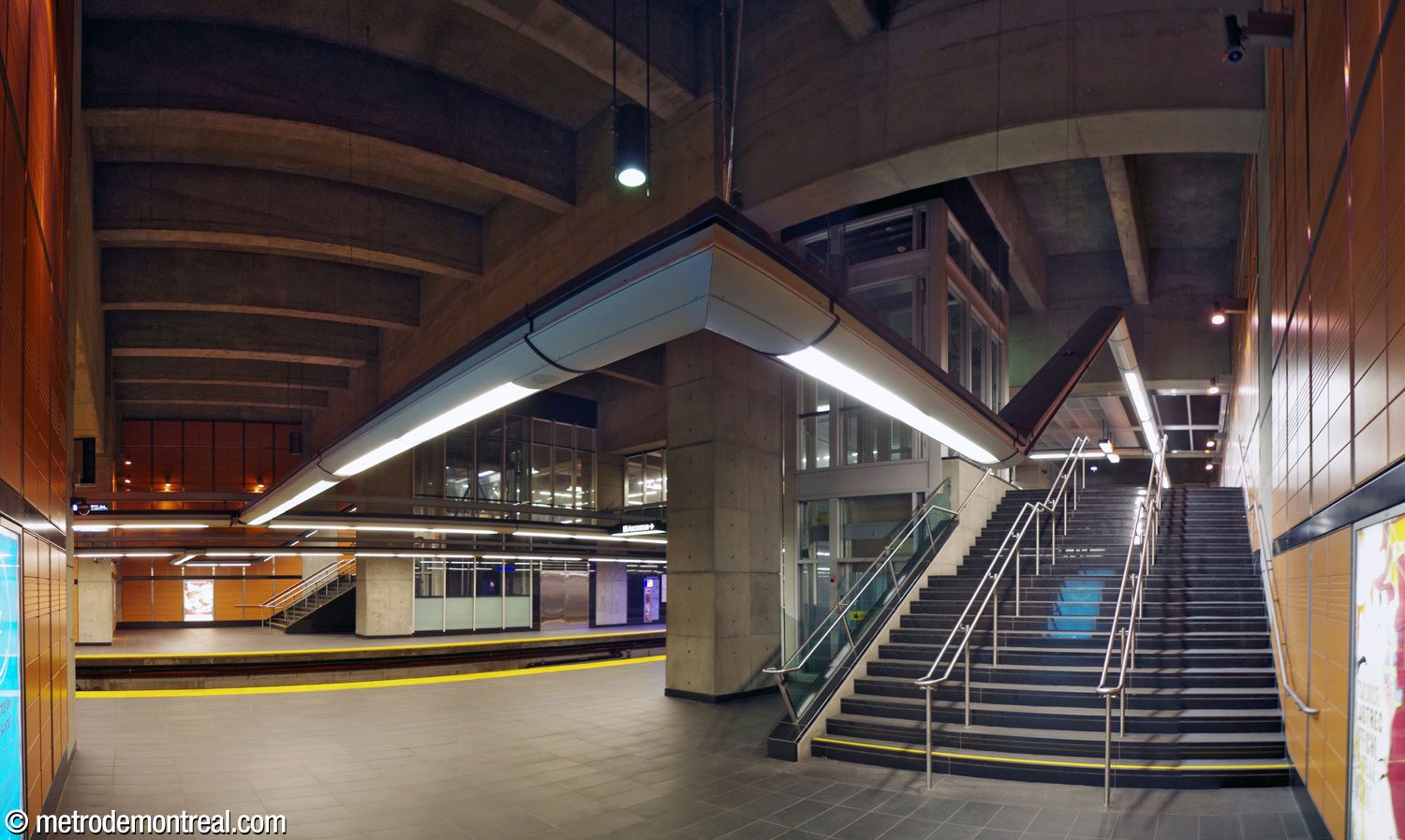 Le pied de l'un des larges escaliers entre le quai Côte-Vertu et la passerelle au niveau de la billetterie. Les éléments d'éclairage suspendus forment des lignes continues qui tracent le parcours des passagers.