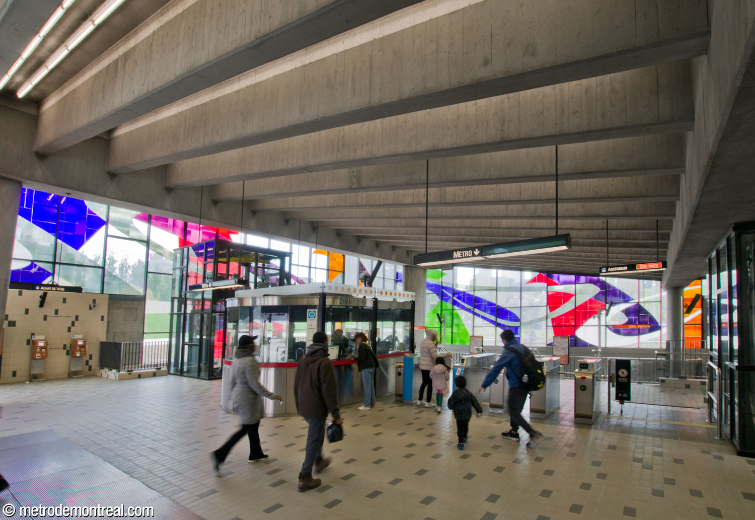 The street level of the headhouse, just inside the main entrance, facing the gateline and ticket booth. The black framework of the elevator shafts rise on either side of the gateline. Marcelle Ferron’s stained-glass window is beyond on the south and west walls.