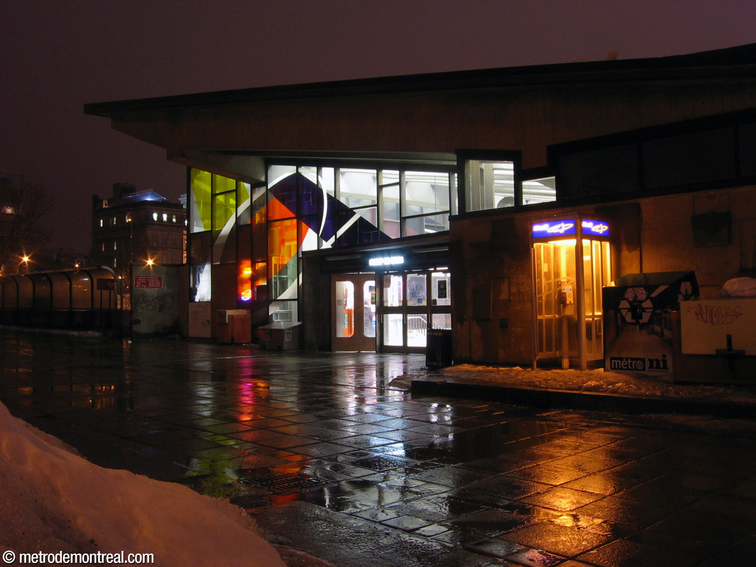 The entrance and eastern wall of the station, showing Marcelle Ferron's stained-glass window illuminated from within by night. (This image was taken before the concession inside the headhouse was moved and before the pedestrian tunnel to Old Montreal and its walkway, left, were demolished.)