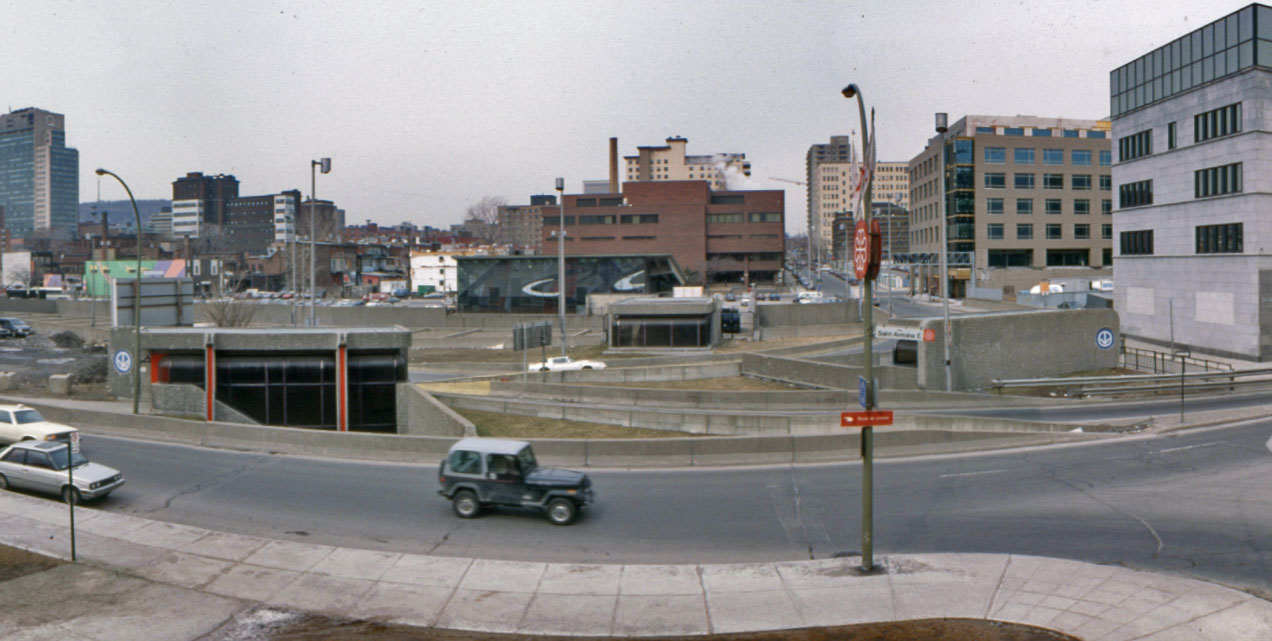 Panorama of the Champ-de-Mars tunnel, seen from the south. The station headhouse is in the centre background; in front of it is the north entrance to the tunnel, at the end of the walkway over the autoroute, next to the headhouse. The two small exits on the north side of Rue Saint-Antoine are at left and right, amid the highway offramps. The large exit in Champ-de-Mars park is behind the viewer.