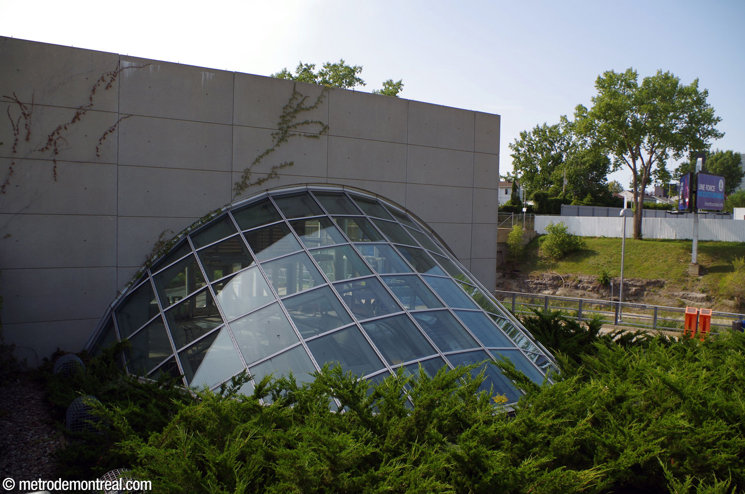 The above-ground, glazed portion of the cylindrical shaft containing the stairs and escalators between the headhouse and ticket hall emerges from the ground behind the headhouse.