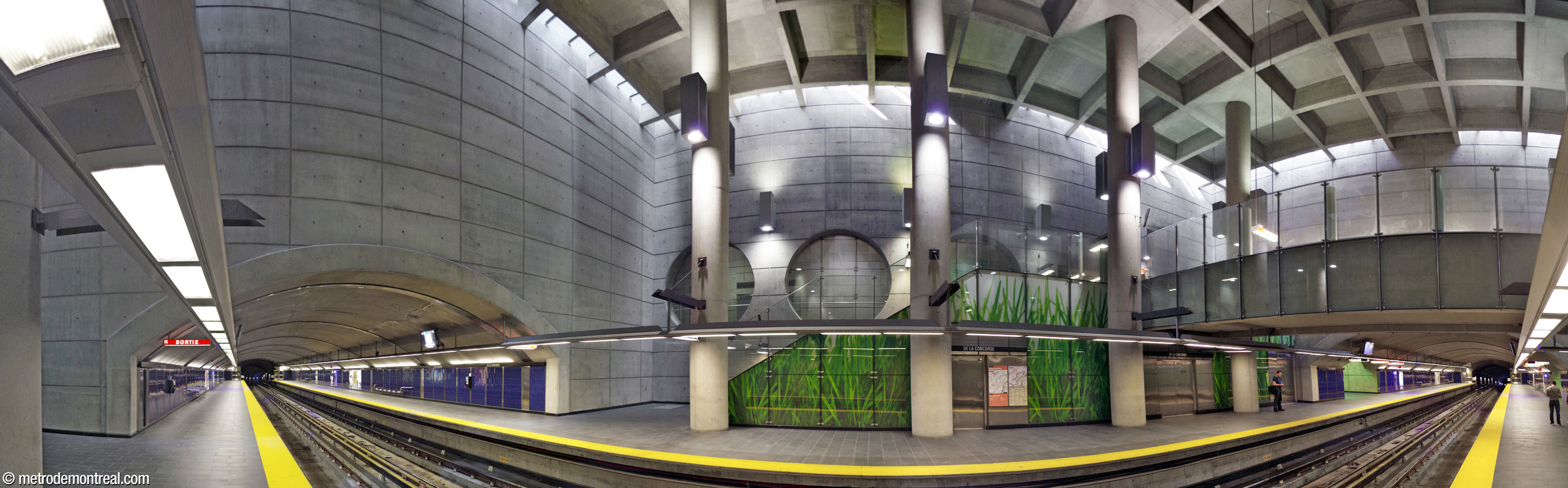 Panorama of the trainroom from the Côte-Vertu platform. The ends of the platforms, built in tunnel, extend out from the large cube-shaped central volume, opening at the bottom of high, flat concrete walls. Tall columns support the coffered ceiling overhead, which is surrounded on all sides by a row of skylights admitting sunlight. Lamps shine up and down from the columns and the walls behind them. At centre, a staircase leads up to the crossover (right); the side of the staircase is decorated with a white and green photograph of greatly magnified blades of grass. Behind and above this staircase is a row of three large circular windows looking onto the hallway between the ticket hall and the elevator to the surface. Nearly all surfaces are in bare concrete, except for the photograph murals and the sapphire blue platform walls.