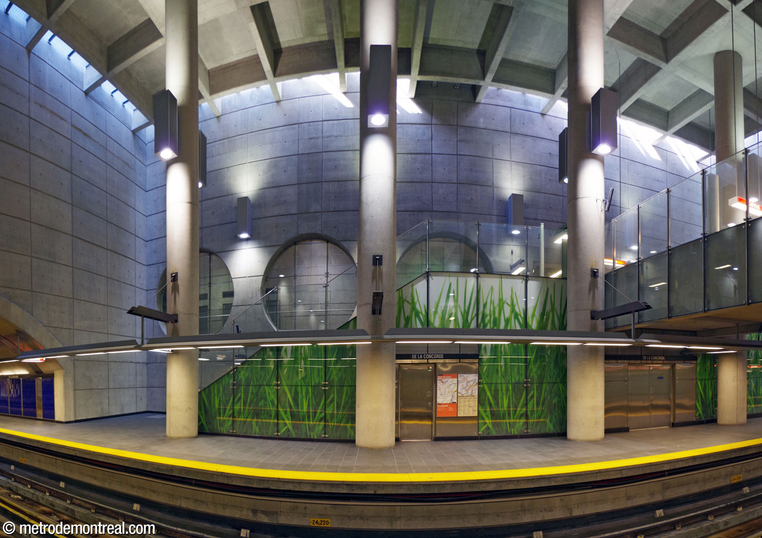 The main staircase from the Montmorency platform up to the crossover level, behind the volume's pillars and in front of the row of circular windows looking onto the hallway to the elevator from the ticket hall to the surface. The wall of the staircase is lined with a large-scale photograph mural of blades of grass.