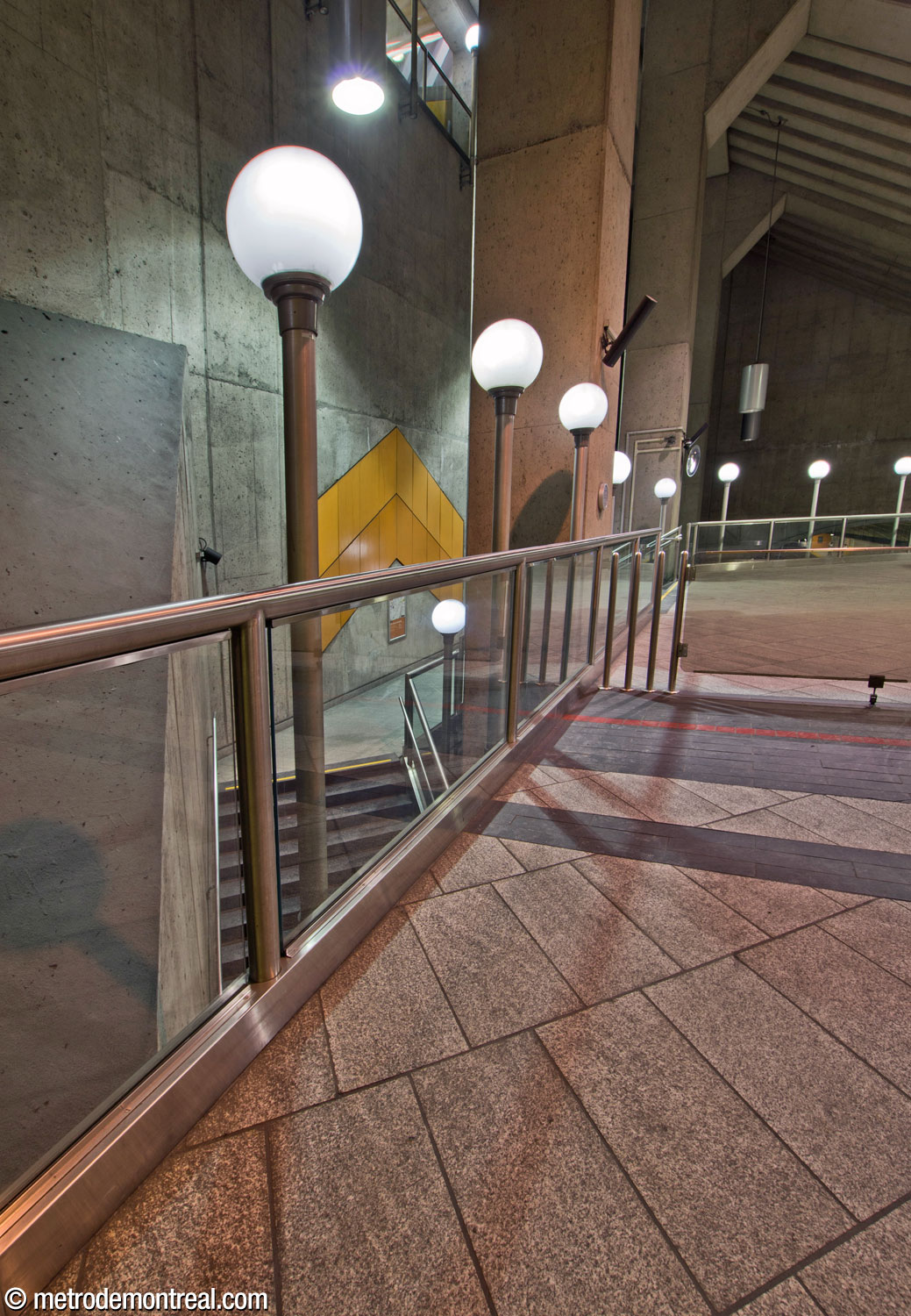 Une autre rangée de lampadaires longe le parapet protégeant le bord est de la mezzanine. En bas, les escaliers vers le quai Côte-Vertu se faufilent entre les colonnes qui appuient la mezzanine et la toiture. Un chevron de Gilbert Sauvé surplombe le palier des escaliers.