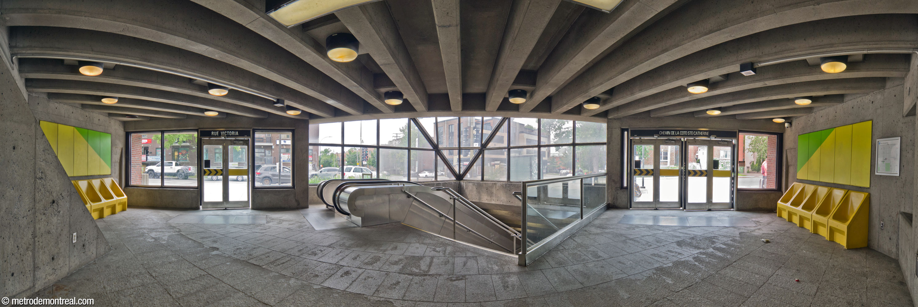 Panorama de l'intérieur de l'édicule, en regardant le long de l’axe de symétrie du bâtiment vers les escaliers et la large baie vitrée au-dessus d’eux. Les sorties donnant sur le chemin de la Côte-Sainte-Catherine (droite) et l'avenue Victoria (gauche) sont disposés de façon symétrique à l’est et au sud de la trémie d’escalier. Deux lignes de bancs du même style que ceux sur les quais sont mises en valeur par des murales de l'architecte.