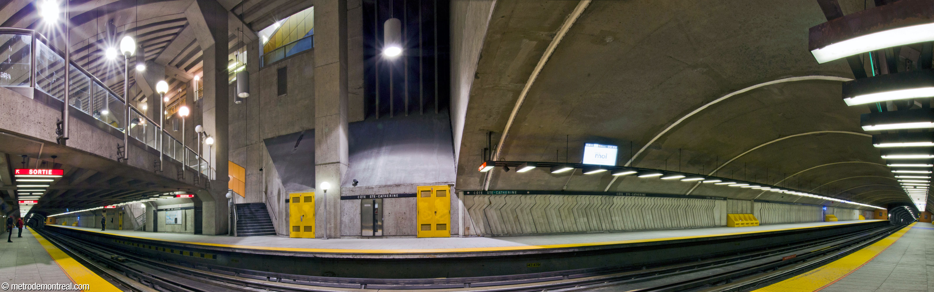 Panorama de la nef de la station Côte-Sainte-Catherine, dont les voûtes excavées en tunnel (droite, extrême gauche) se projettent du volume central (centre gauche). Le toit du grand volume est incliné vers le haut d’ouest en est (en haut à gauche), et son faîte s’ouvre sur un passage qui le surplombe. Le parapet à la fin de la mezzanine traverse les quais en diagonale. Au centre, une bouche de ventilation est visible à l’extrémité sud du volume.