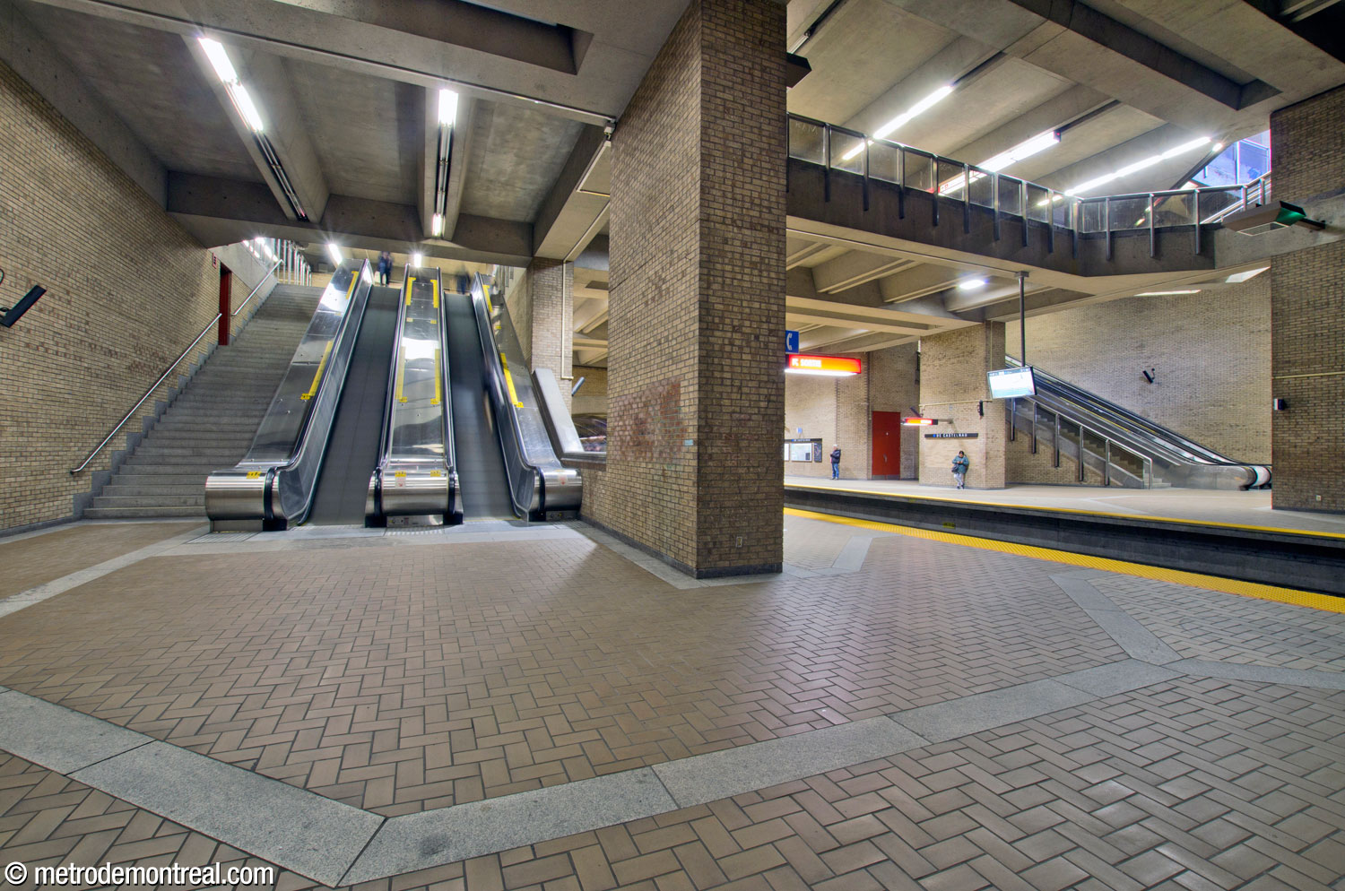 The side aisle of the Snowdon platform, leading to the stairwell up to the ticket hall. A row of paving stones leads from the stairs to the platform. The mezzanine level is visible above the main volume.