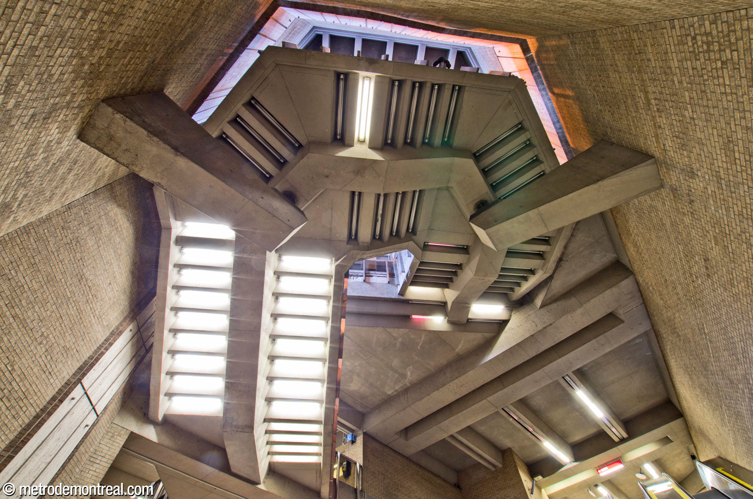 The U-shaped staircase between the Saint-Laurent Ouest headhouse and the ticket hall, seen from the Saint-Michel platform below. The monumental staircase is separated from the adjacent walls and seems to hover in the lofty volume, supported by large corbels. Fluorescent lamps on its underside illuminate the volume, along with natural light that filters down from the headhouse above.