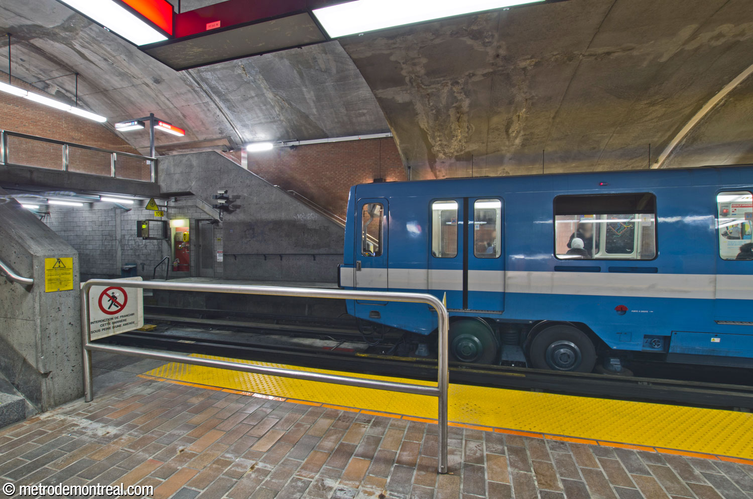 A Saint-Michel-bound train stops at the eastern end of the platform, at the stairs leading up to the balcony-style crossover at that end. In the foreground, a barrier on the opposite platform reveals the small offset between the standing positions of Snowdon- and Saint-Michel-bound trains.