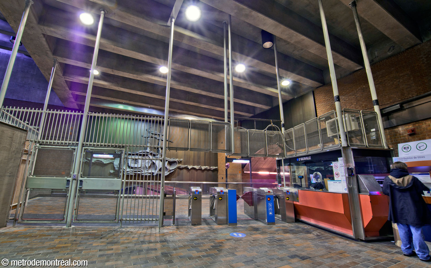 The unstaffed gateline of the Louis-Hébert access, facing a parapet overlooking the platforms. Through the fence surrounding the gateline, Eddy Tardif's sculpture Le Pélican is visible on the spandrel wall over the trainroom vault.