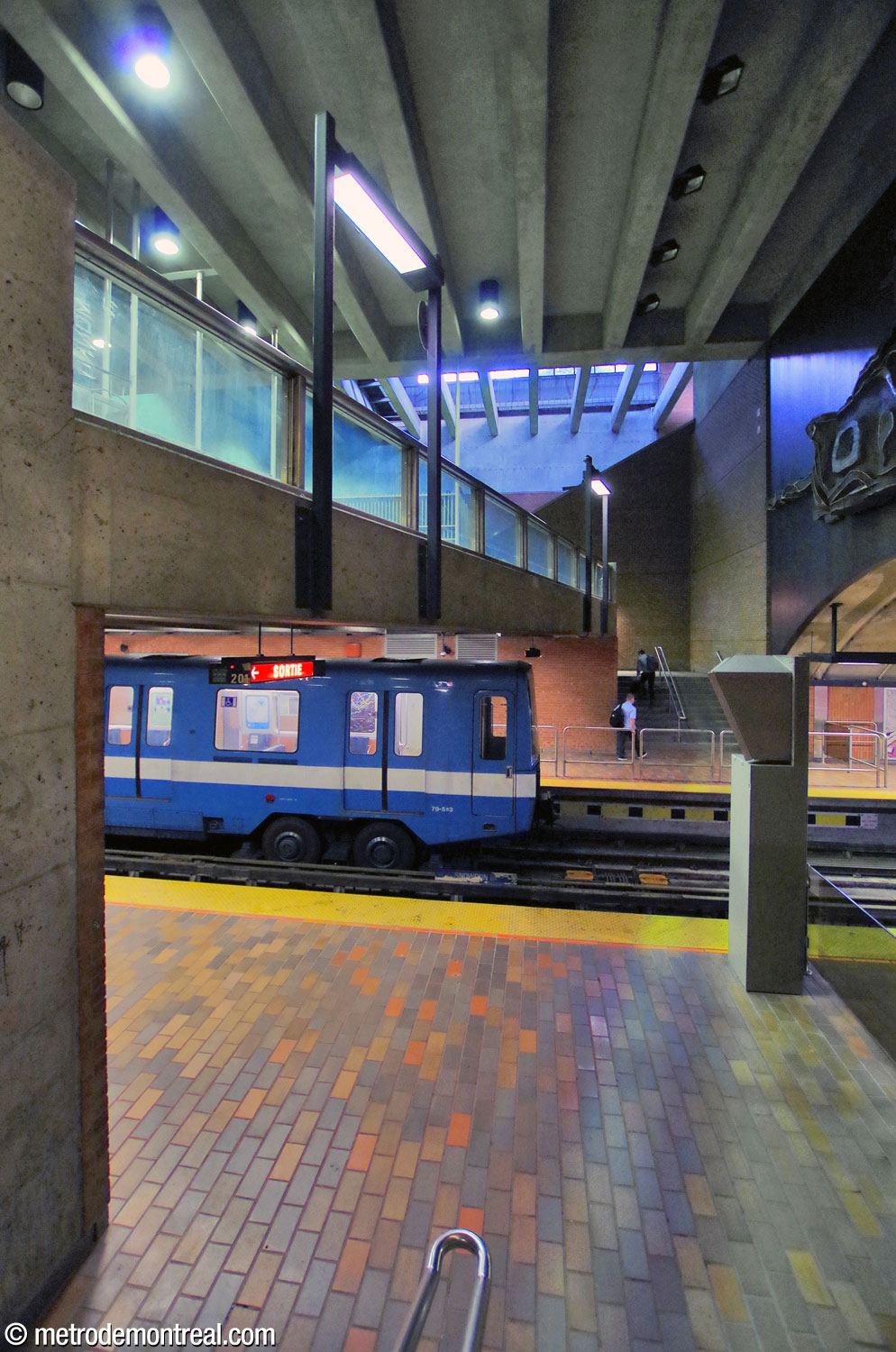 The tall volume of the Louis-Hébert access above the platforms, seen from the stairs to the Snowdon platform. In the background, natural light from the headhouse illuminates the escalators to the exit. The parapet of the ticket hall mezzanine passes diagonally over the tracks. The tail of the Saint-Michel-bound train standing at the opposite platform shows the offset of the platform faces.