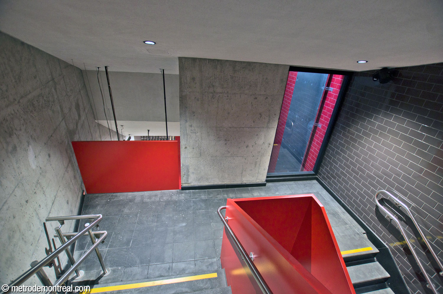 L’escalier auxiliaire accompagnant l’ascenseur principal est encadré par une balustrade et un parapet écarlate vif et un mur d’accent noir. On regarde vers le deuxième palier sous le rez-de-chaussée, où un parapet donne sur le niveau du transept en contrebas et une fenêtre laisse voir l’intérieur de la gaine d’ascenseur.