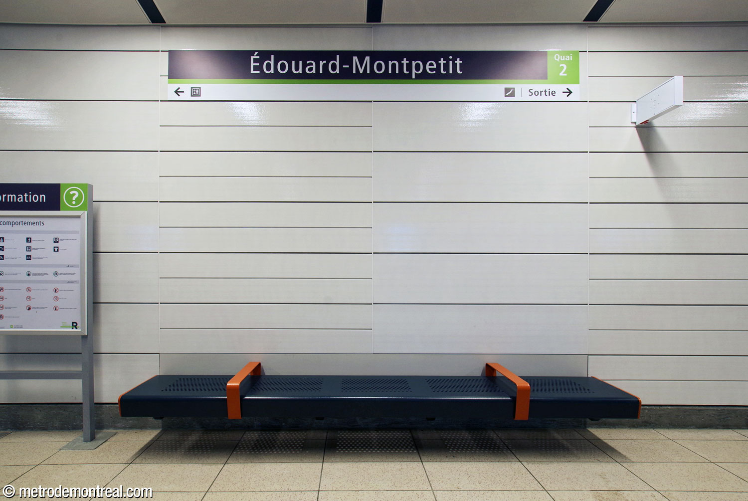 Un banc du quai, en métal noir avec des appuis rouges en conformité avec la couleur thématique de ce tronçon du REM. Derrière lui, le revêtement mural métallique émaillé blanc, utilisé dans toute la station.