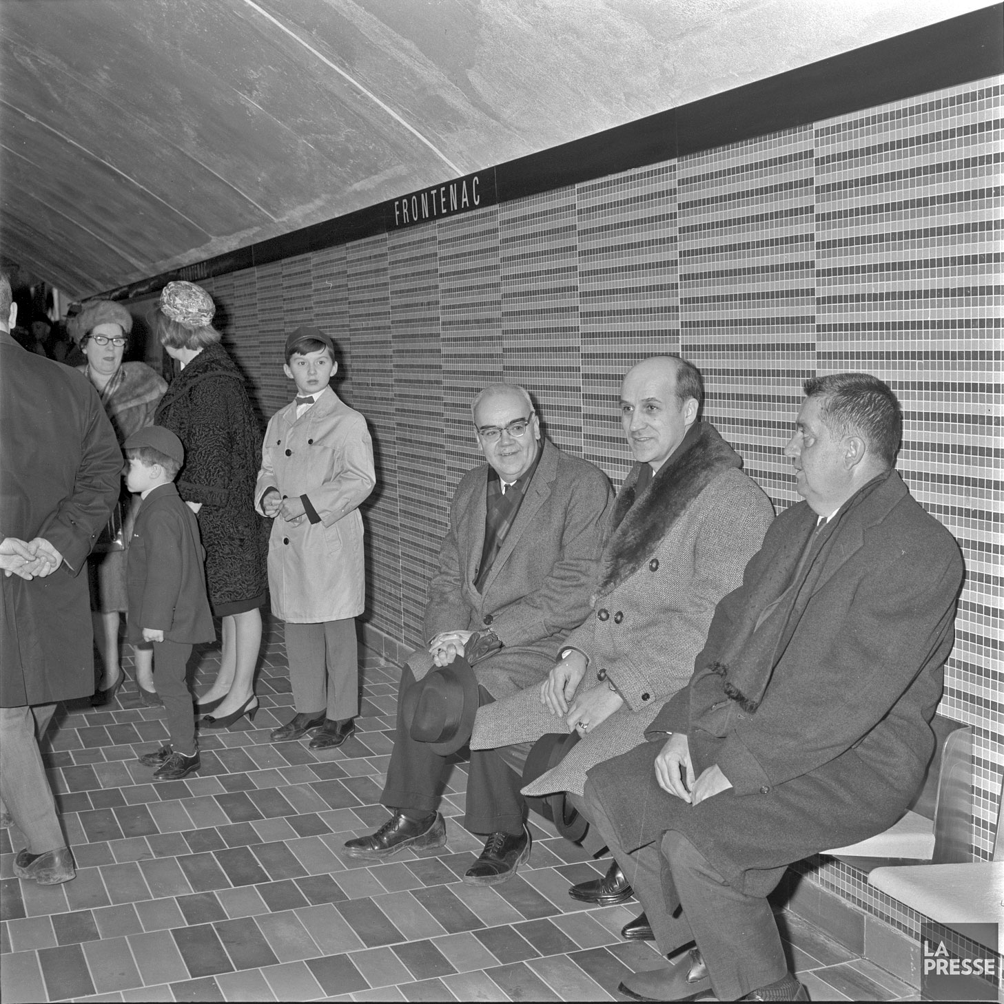 Les dignitaires sur le quai durant la cérémonie d’inauguration de la station le 18 décembre 1966. Sur le banc, de gauche à droite : Lucien L’Allier, président de la Commission de transport de Montréal; Paul-Émile Sauvageau, député provincial de Bourget; et Maurice Allard, président de l’Association des hommes d’affaires du quartier Frontenac. À noter, le motif originel du carrelage mural derrière eux.