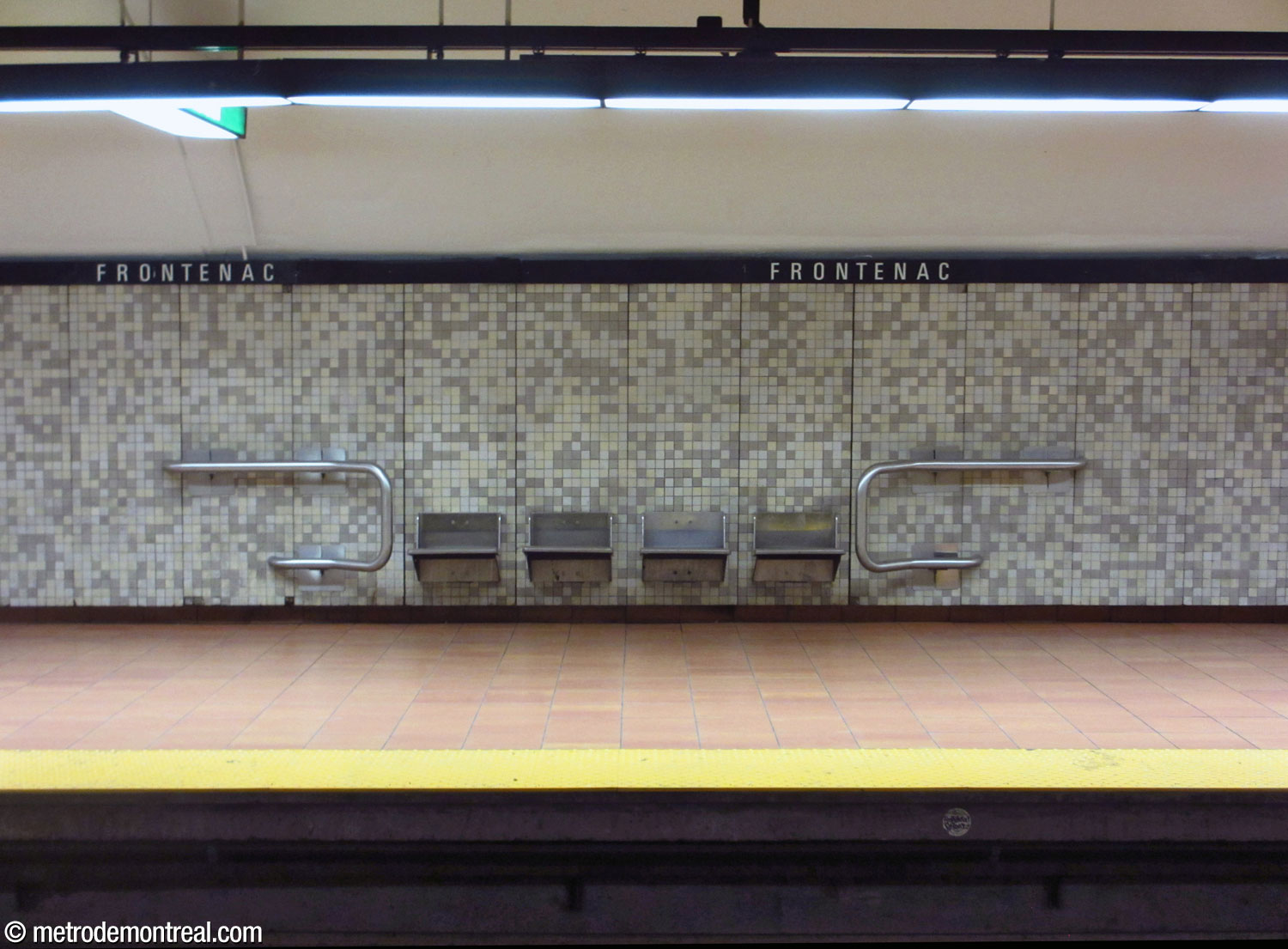 Un banc de sièges en acier inoxydable au niveau du quai, avec le carrelage gris et beige qui caractérise l'ensemble de la station.