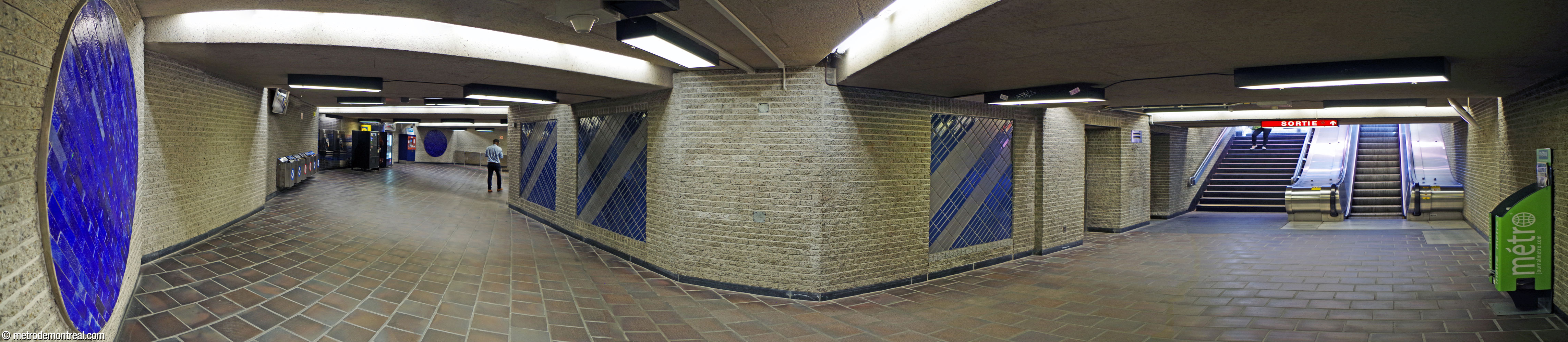 Panorama du niveau de la billetterie. Le corridor à partir des tourniquets (gauche) prend un virage pour atteindre l'édicule (droite). Les murs de brique de béton sont enjolivés de cercles bleus et de panneaux de carrelage bleu et gris.