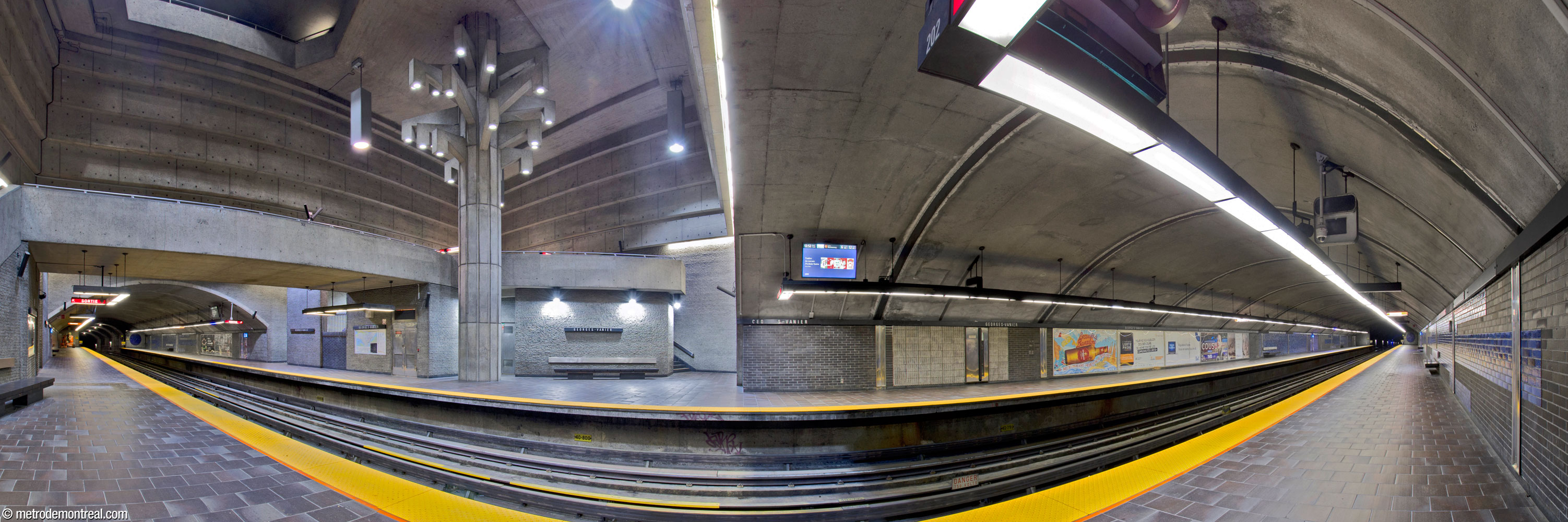 La nef de la station Georges-Vanier. Creusée en tunnel, elle projette d’un volume central, qui accueille une section élargie de chaque quai. La sculpture Un Arbre dans le parc se dresse dans le volume au-dessus de la section élargie du quai Côte-Vertu, devant la passerelle (gauche). Le volume est revêtu d’un lambris en brique de béton sous une partie supérieure en béton brut; les portions des quais construites en tunnel sont parées de brique noir et bleu saphir.
