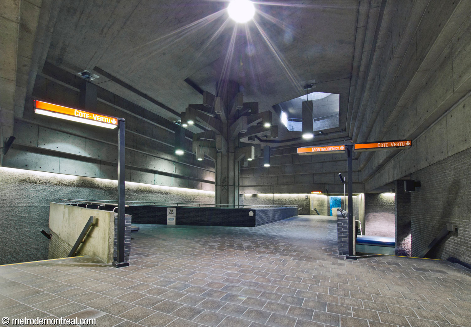 Le volume central de la station, vu à partir du pied des escaliers vers la sortie. Des escaliers descendent vers le quai Côte-Vertu de part et d'autre de la passerelle vers le quai Montmorency. À l'extrémité proche, la passerelle s'élargit pour former une aire d'attente avec un banc. La sculpture Un Arbre dans le parc surplombe la passerelle. Un puits d'éclairage dans le plafond laisse jaillir le soleil.