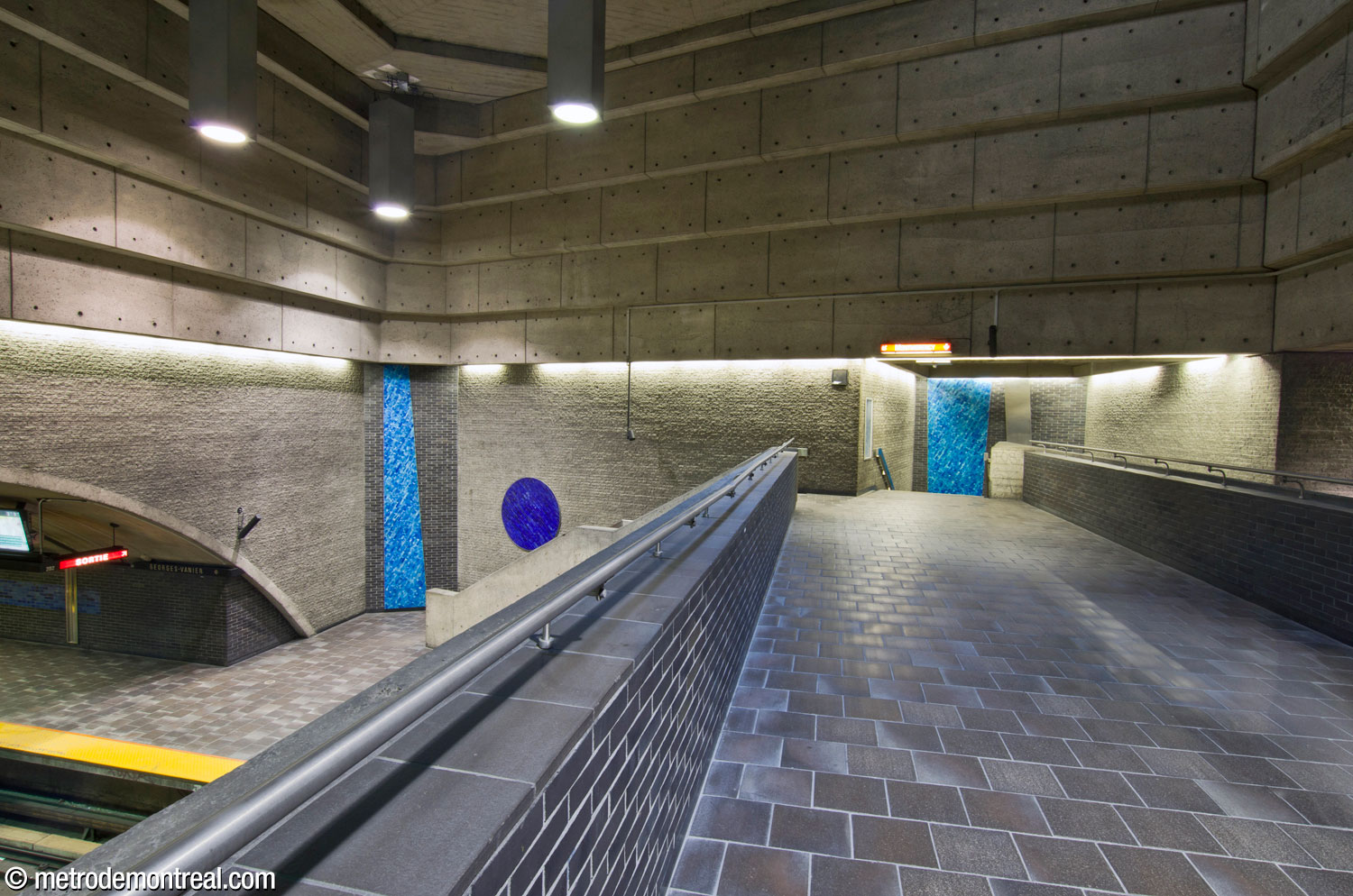 Vue à partir de la passerelle vers le sud et les escaliers reliant ce niveau avec le quai Montmorency. Les murs du volume et des cages d'escaliers sont revêtus de trapèzes carrelés couleur turquoise et d'un cercle bleu saphir au-dessus de l'escalier à gauche.