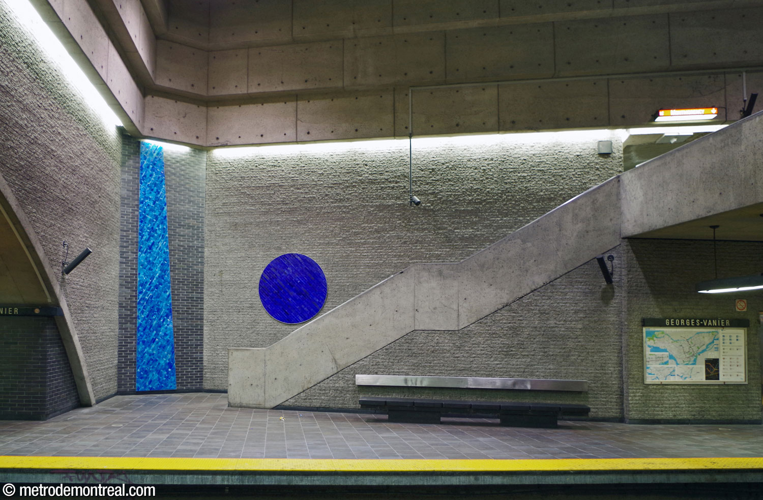 L’escalier du côté est de la passerelle qui la relie au quai Montmorency, dans le volume central. L'escalier et le mur adjacent sont disposés à un angle oblique relativement à la passerelle et le bord du quai. Un cercle carrelé bleu saphir et un trapèze turquoise égaient le mur de brique gris pâle.