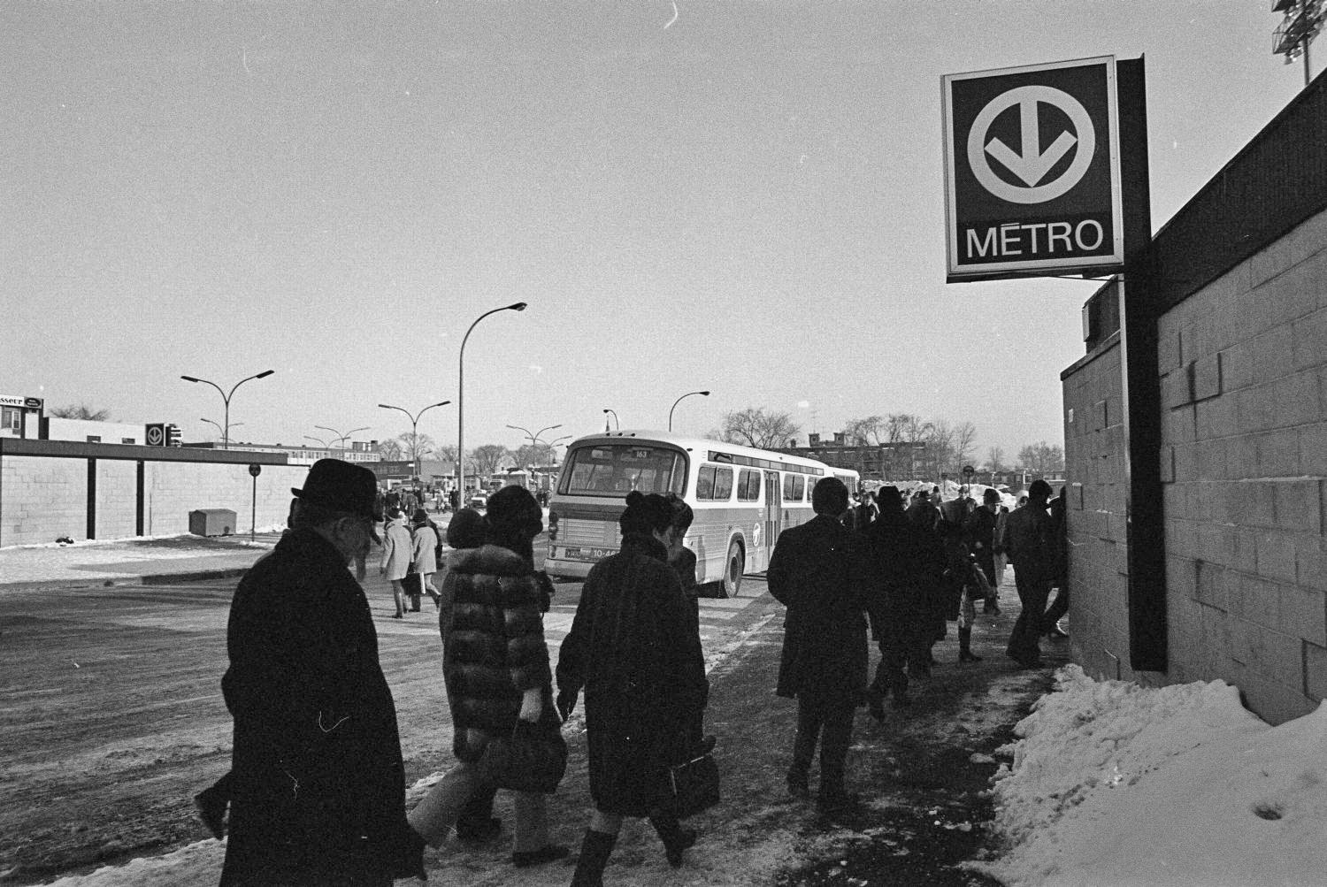 Des passagers attendent leur autobus à côté de l'édicule sud du terminus d'autobus; on voit également l'édicule nord, à gauche.