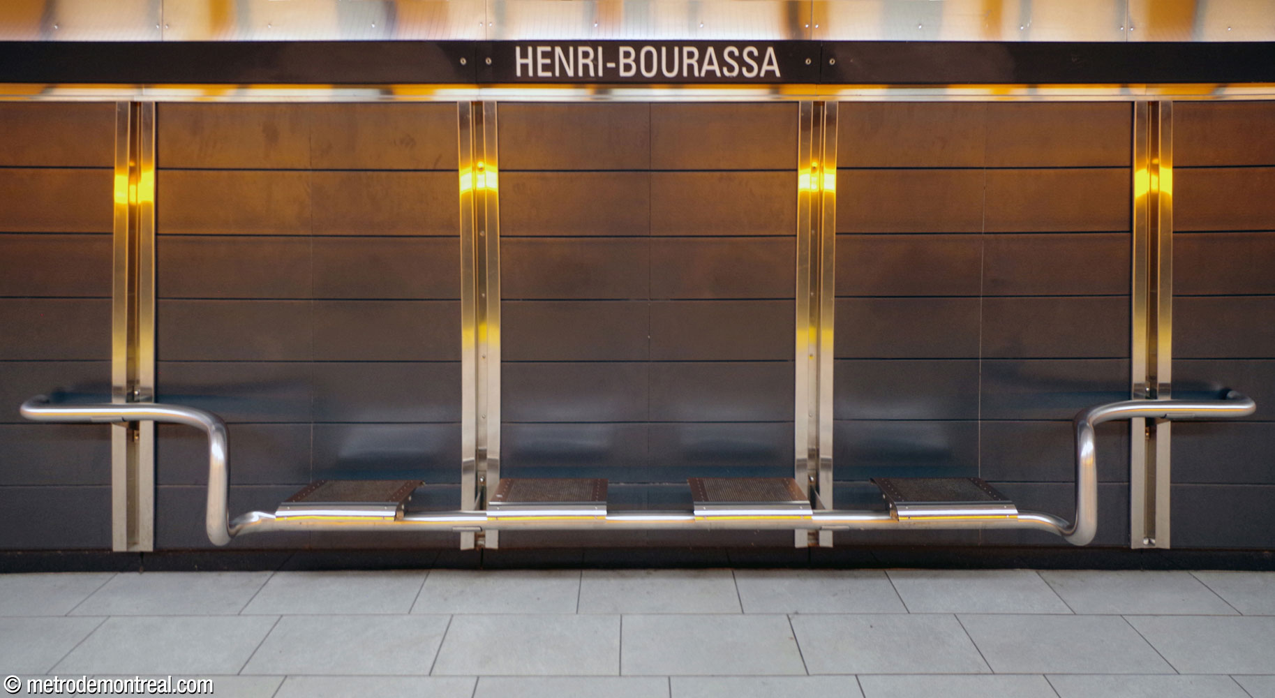 Un banc en acier inoxydable sur le quai Montmorency, sur le fond de panneaux gris ardoise et les jointures également en acier inoxydable. On voit aussi les reflets de la bande lumineuse orange au-dessus de la voie.