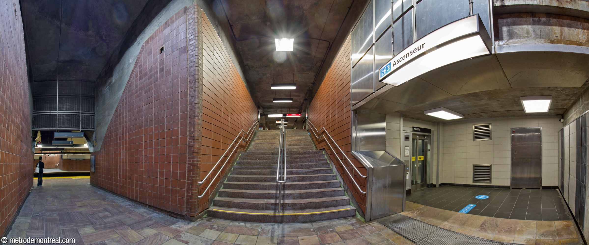 L’escalier nord vers le quai Côte-Vertu. Chacun des quatre cages d’escalier vers la nef originelle passe par un coin de 90 degrés pour atteindre le quai. Les deux cages d’escalier nord descendent jusqu’au niveau du quai avant de tourner. Le passage entre le pied de l’escalier et le quai fait face à son homologue de l’autre côté, et les deux constituent une boîte qui entrecoupe la voûte du tunnel. La partie supérieure de l’embouchure est désormais fermée avec un grillage, sans doute à des fins de sécurité incendie. À droite, le pied de l’ascenseur vers le niveau de la passerelle.