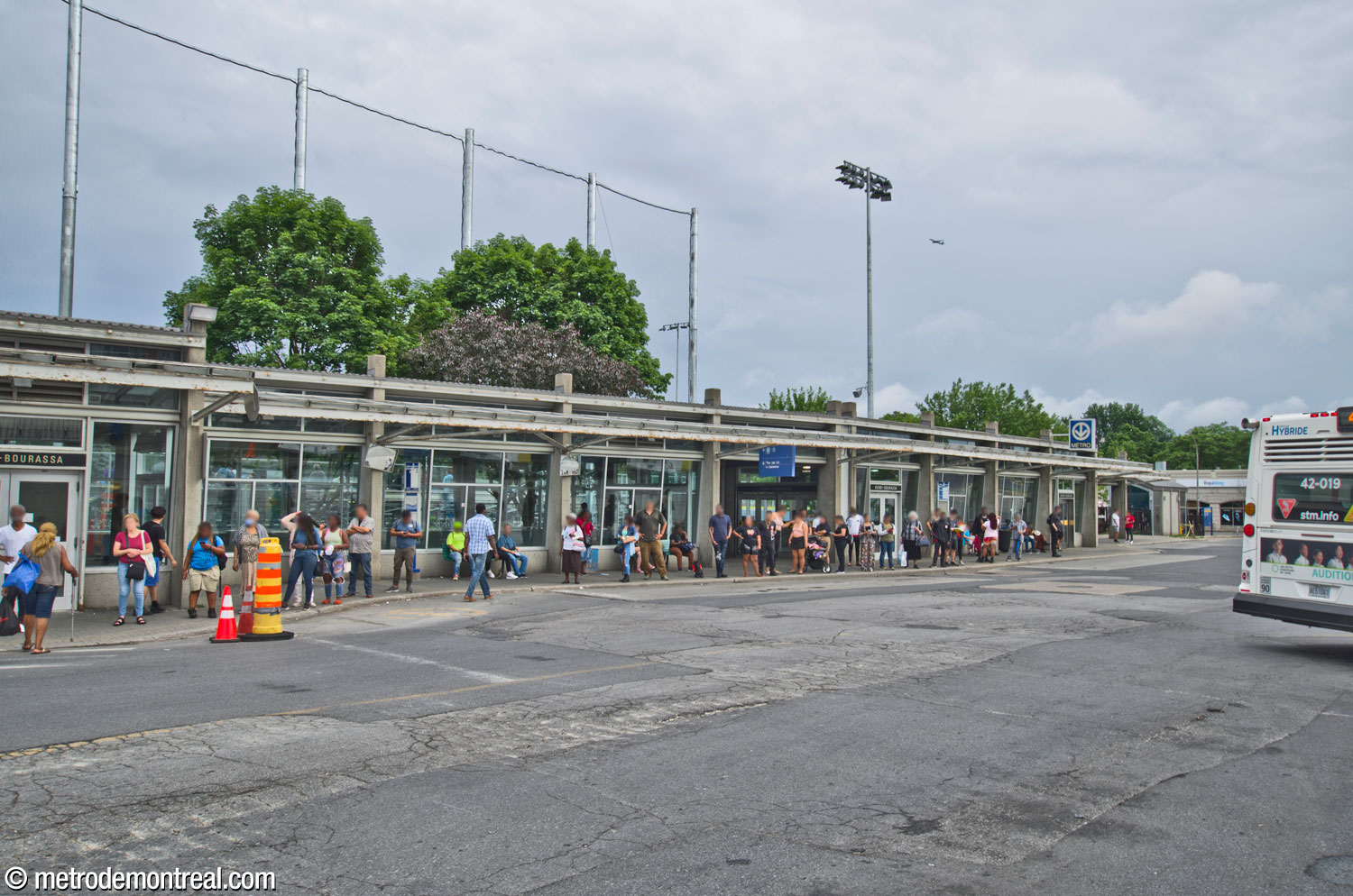 Une foule attend des autobus à l’extérieur de l’îlot sud du terminus d’autobus Henri-Bourassa Sud, vu depuis l’îlot nord. La portion du bâtiment à droite contient l’accès vers la station; elle est séparée de l’autre portion du bâtiment à l’est par une courte section recouverte mais ouverte sur le devant.