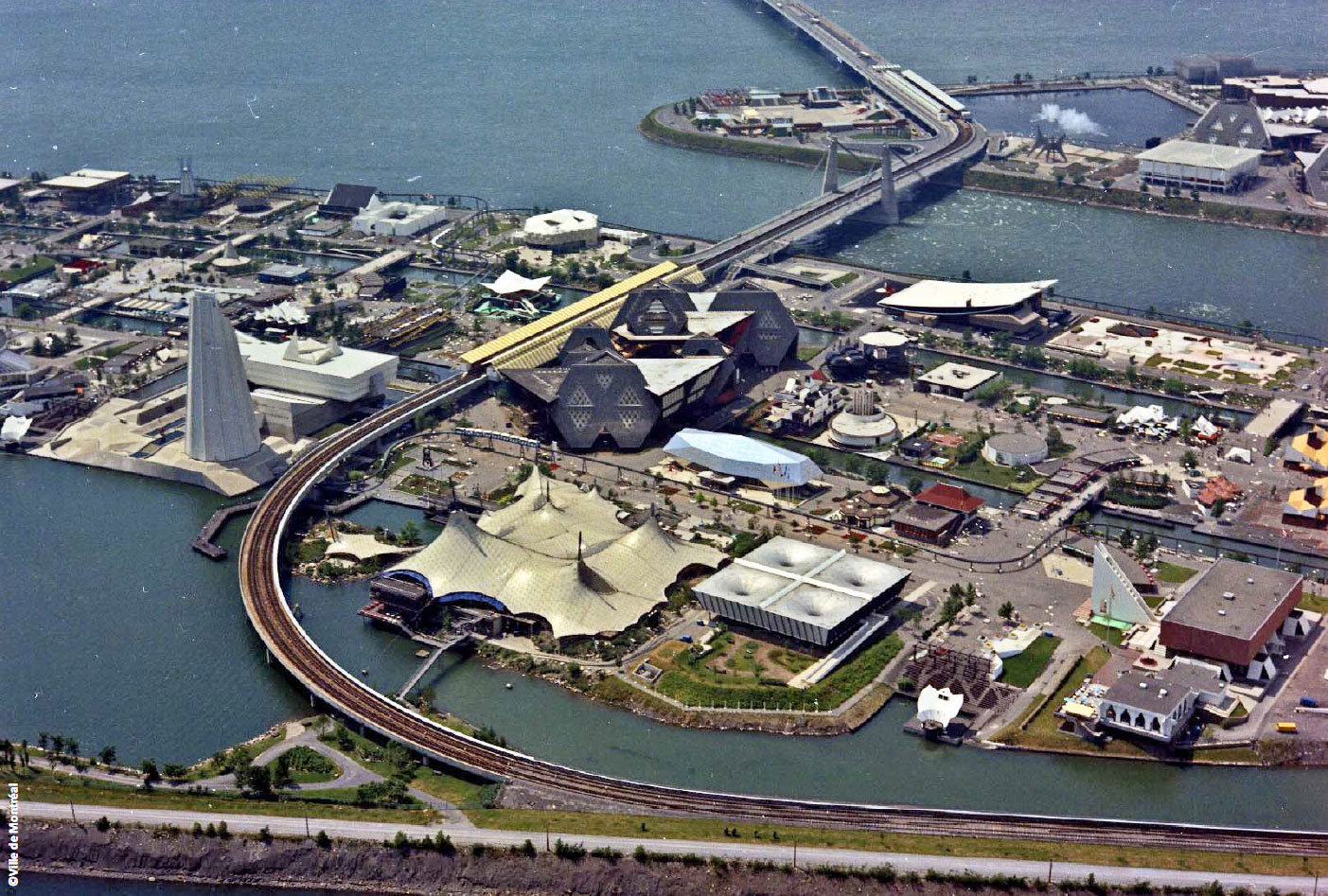 Vue aérienne montrant une partie de la route de l’Expo-Express. La station Île Notre-Dame, au toit jaune, est au centre; au fond, à travers le chenal Le Moyne, on aperçoit la station Île Sainte-Hélène (Place des Nations).