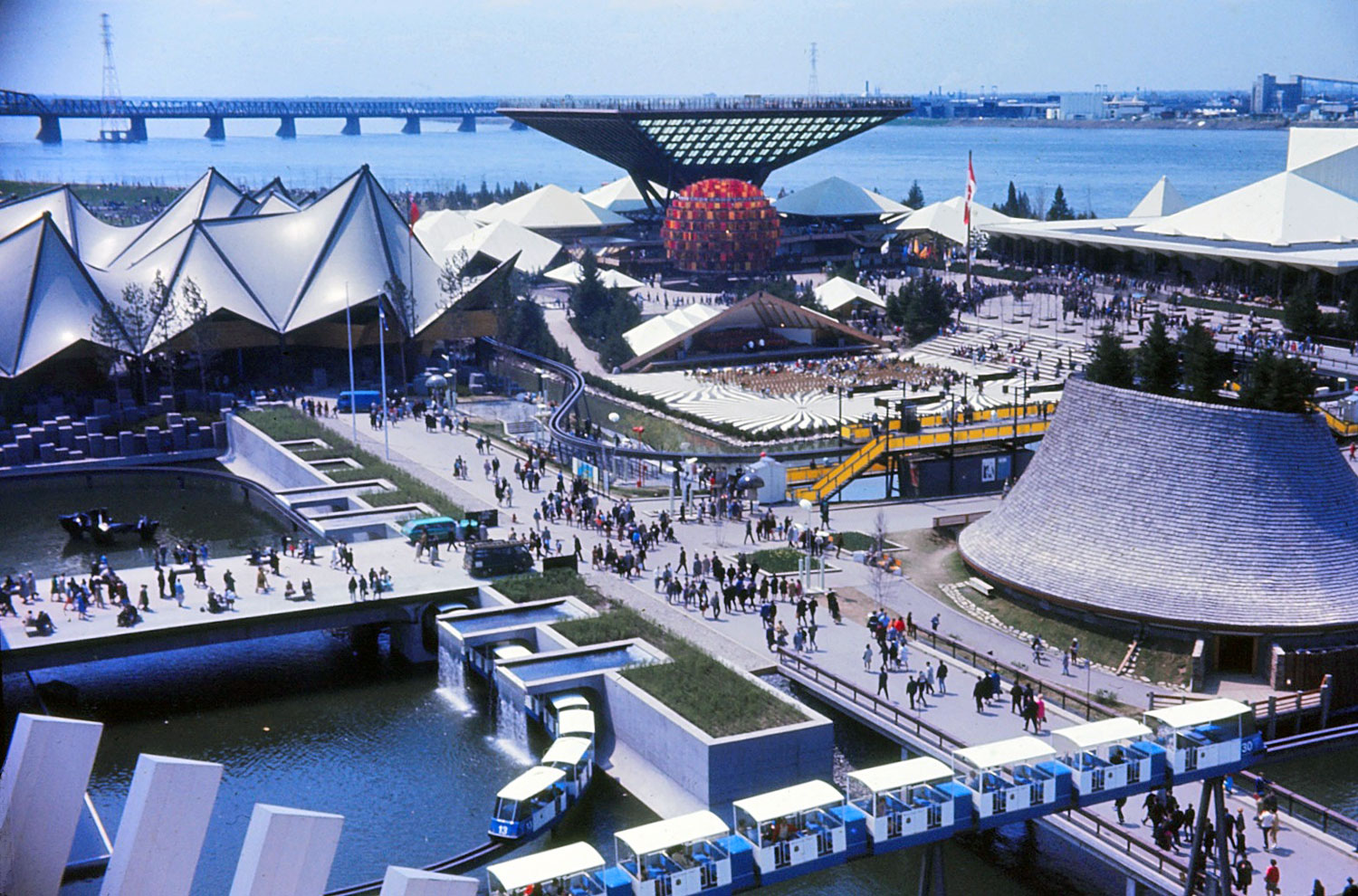 Le système de monorail léger Minirail se fraie un chemin serpentin parmi les pavillons de l’île Notre-Dame. La station Canada, en jaune, apparaît au centre droit; à sa gauche, la voie pénètre le pavillon de l’Ontario. À gauche de l’image, elle ressort pour border le bassin autour du pavillon du Québec, puis se lance haut dans les airs pour contourner le pavillon français. On voit deux trains sur les voies en avant-plan.