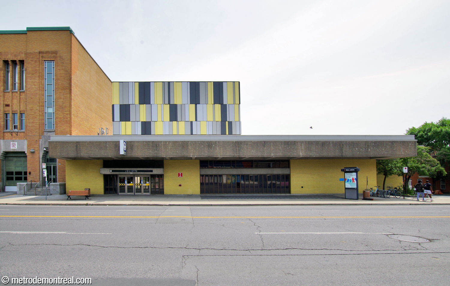 La façade antérieure de l’édicule sud, vue à travers la rue Hochelaga. Ce local technique plutôt lourdaud, revêtu de panneaux jaunes et gris pâle et foncé, est juché maladroitement sur le toit.