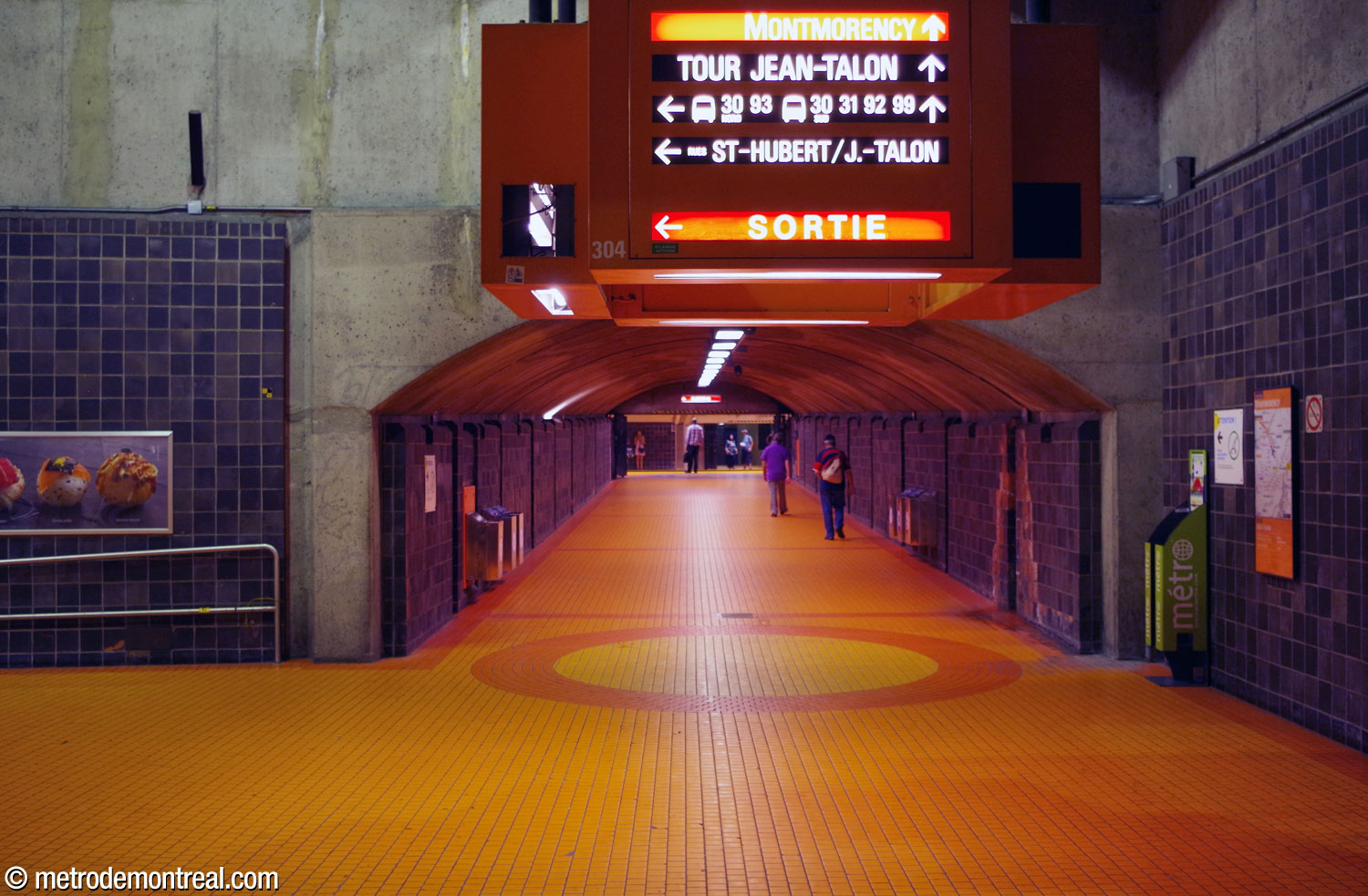 Le tunnel entre la galerie est et le quai Montmorency. Le niveau de la station est indiqué par le carrelage orange au sol et le porte-enseigne orange, alors que la direction du lien est indiquée par la rosace orange.